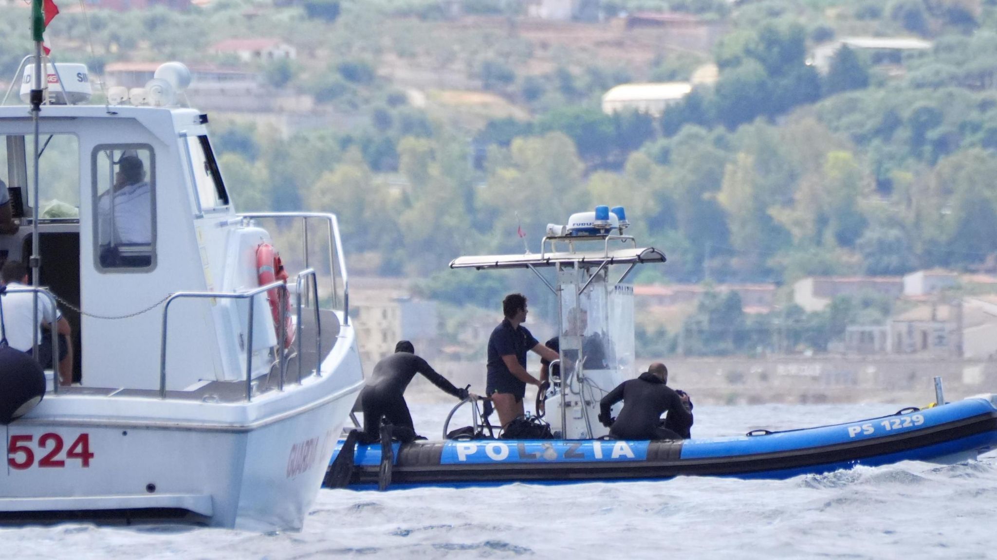 Police divers on a rigid inflatable boat, with a larger boat in the foreground. They are part of the recovery operation above the wreck of the luxury yacht, Bayesian.