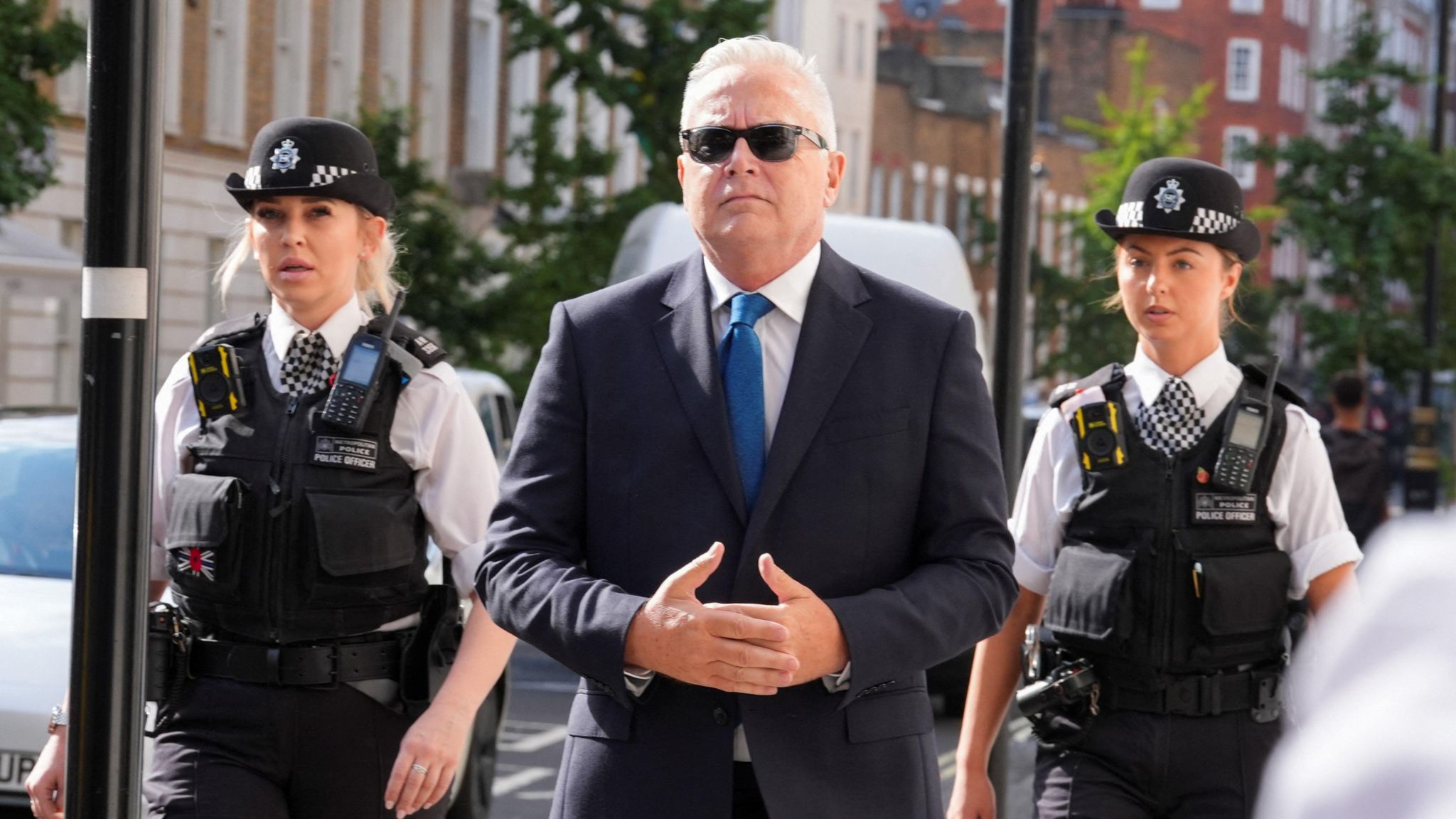 Huw Edwards, an older man wearing a suit, blue tie and glasses walking with two policewoman at either side of him