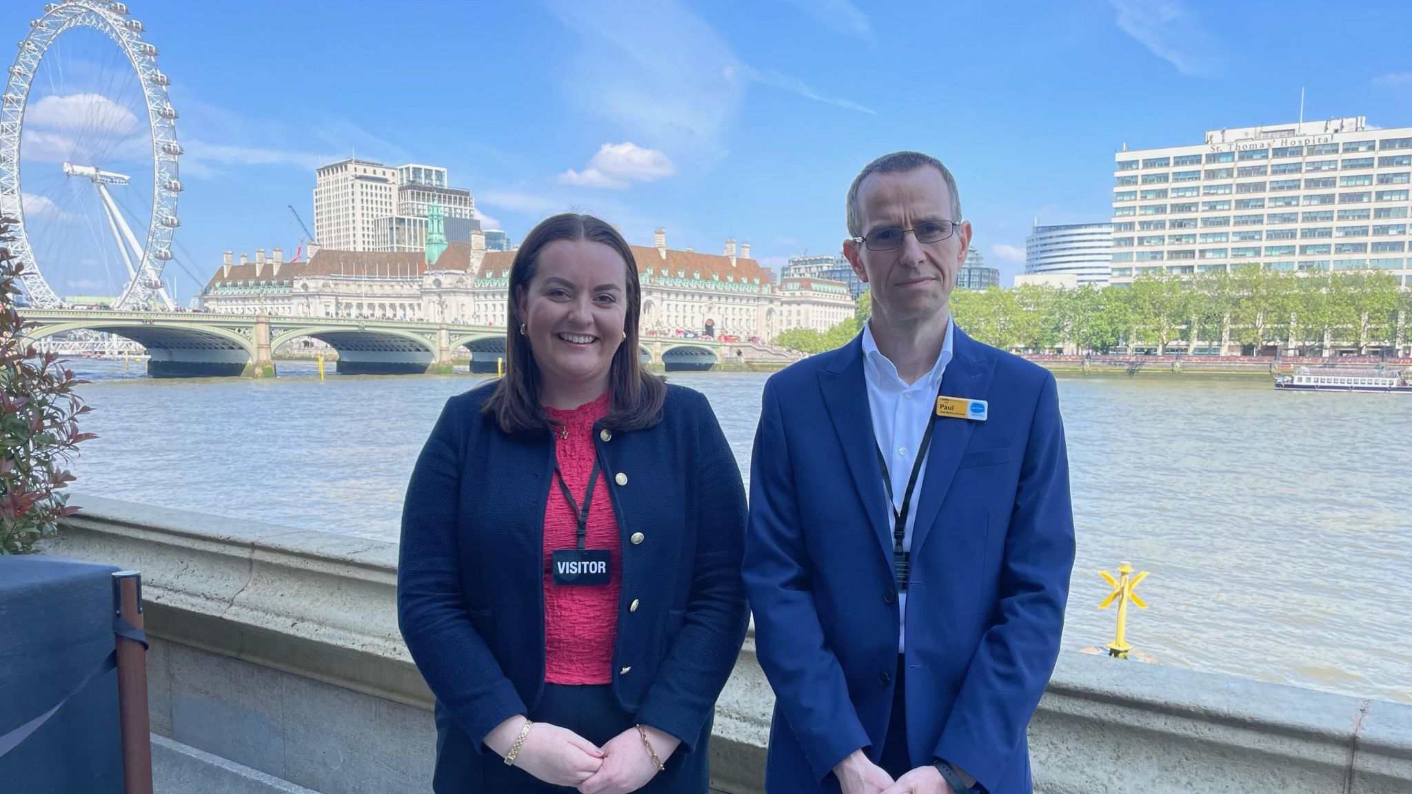 Naomi Barlow, standing in a dark blue suit and pink shirt, with Dr Paul Perkins - wearing a dark blue suit - stood next to her in front of the River Thames in front of the Houses of Parliament