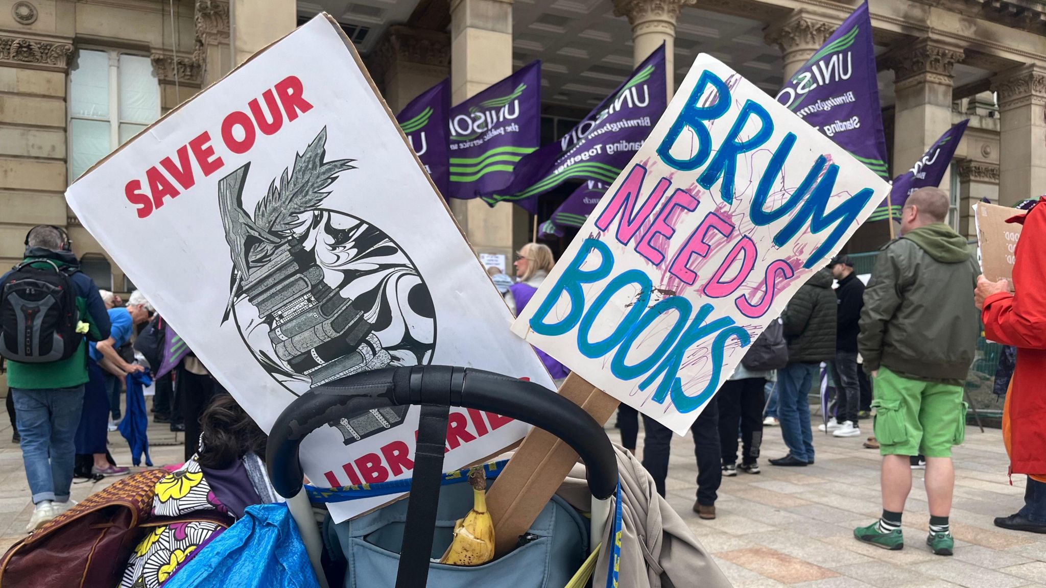 Save our libraries signs in a bag, with protesters in the background demonstrating against cuts to services in Birmingham