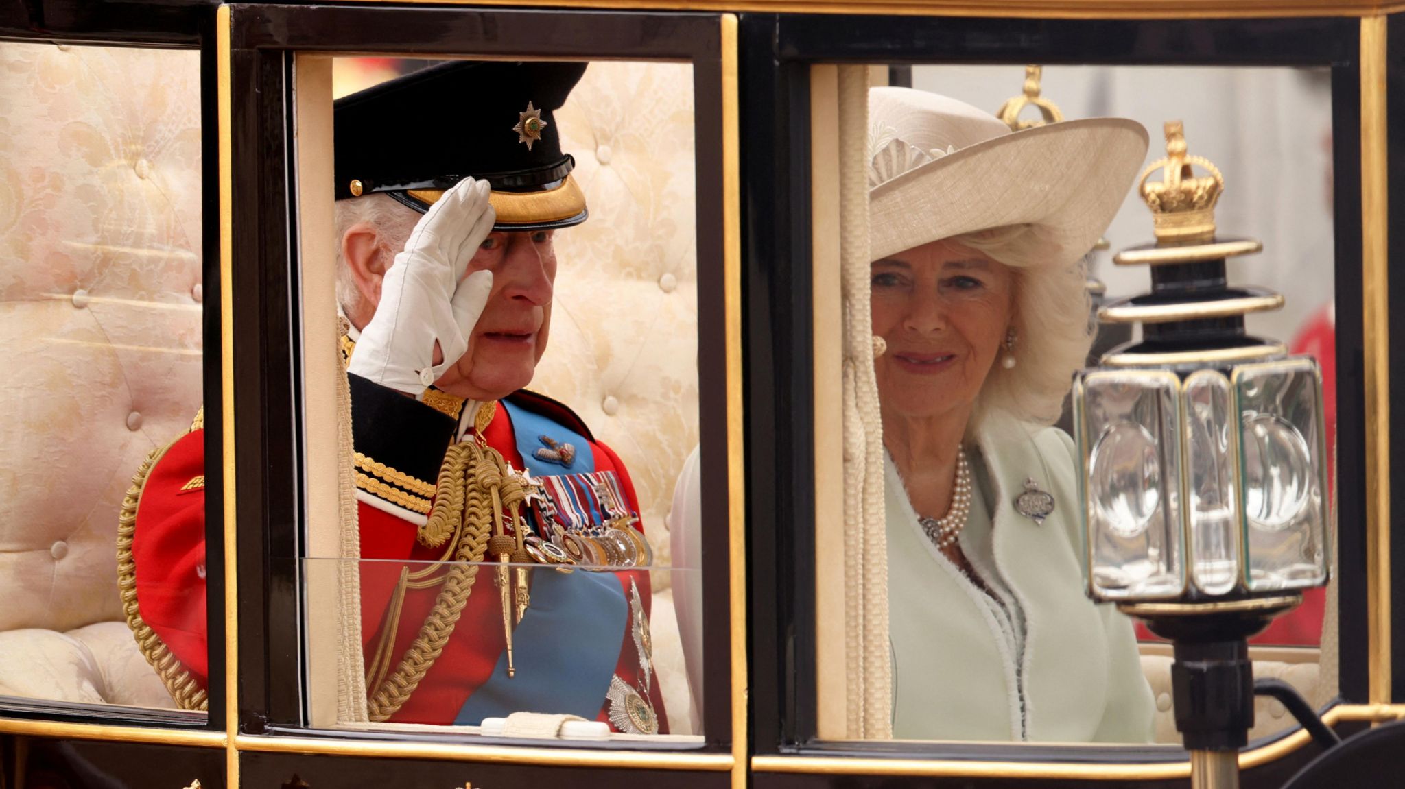 The King salutes from inside his carriage while sitting next to the Queen