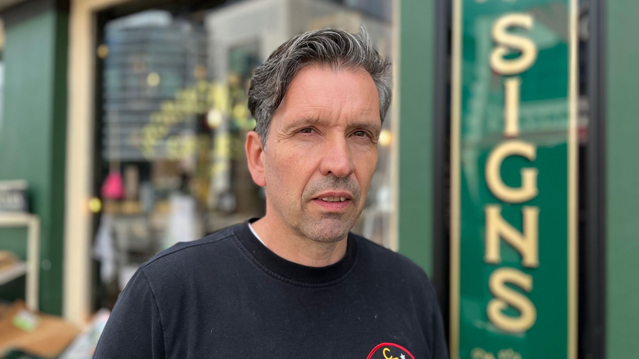 A middle aged white man with black and grey hair and wearing a black tshirt stands in front of the green shop front looking at the camera