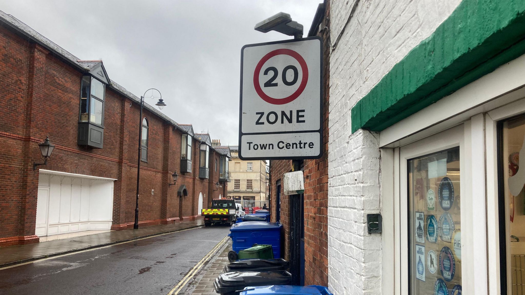 A street sign with a 20 mph speed limit, next to a wall with bins lined up next to it