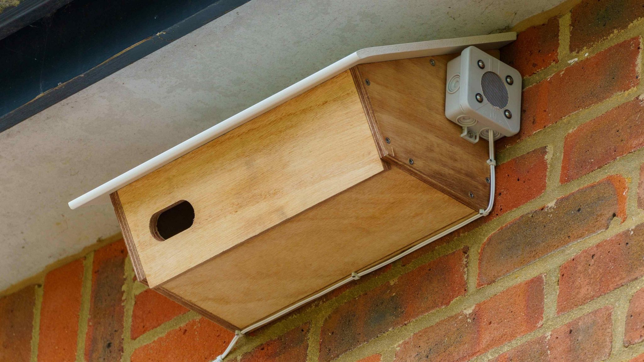 A wooden swift nesting box on the side of a house with a small speaker attached at one end