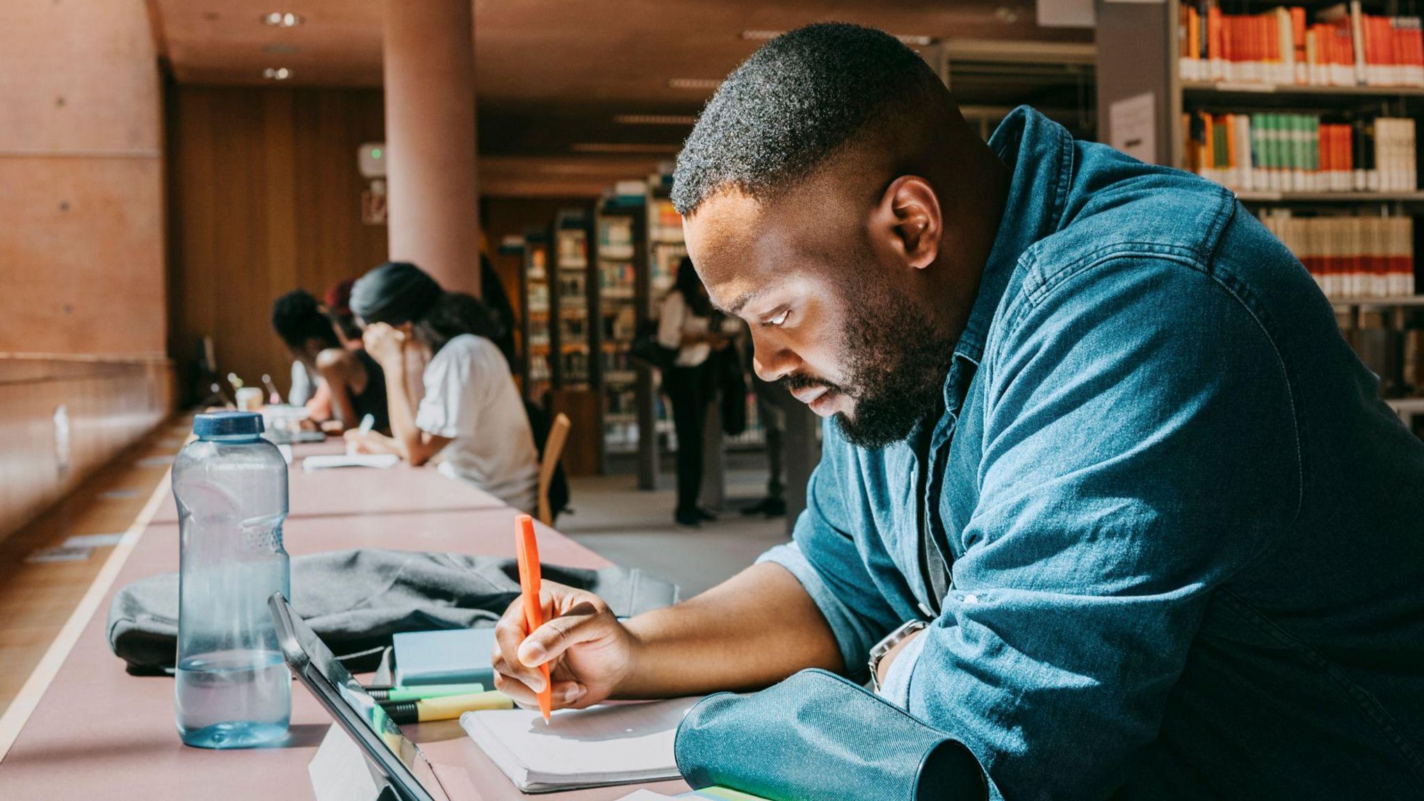 A student with black hair and a black beard sits at a desk in a university library, with bookshelves in the background. He wears a denim shirt and is writing notes with an orange pen, with his laptop screen and a clear water bottle in front of him.