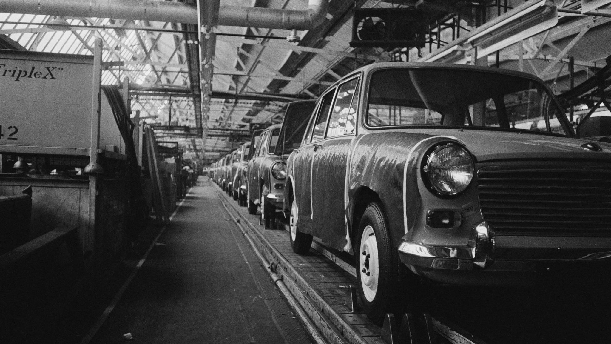 A black and white picture of the production line at Cowley in 1965, with nearly finished cars waiting to roll off it