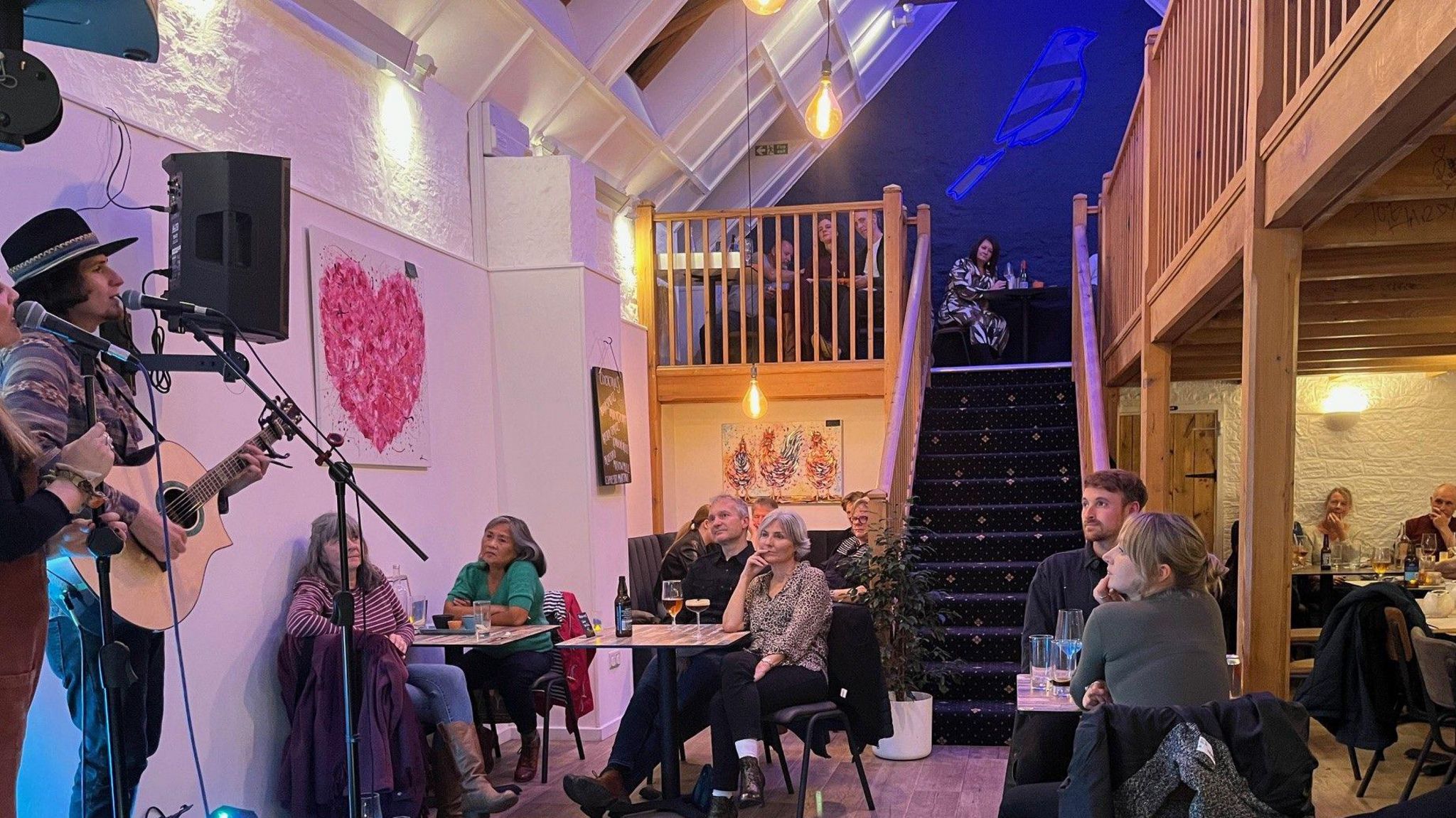 People sit at tables and watch local artist play guitar on stage in the cafe music venue