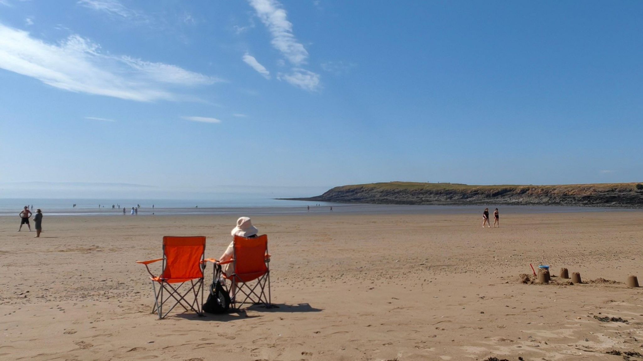 A wide, sunny beach with someone in a deck chair and a sandcastle in the foreground