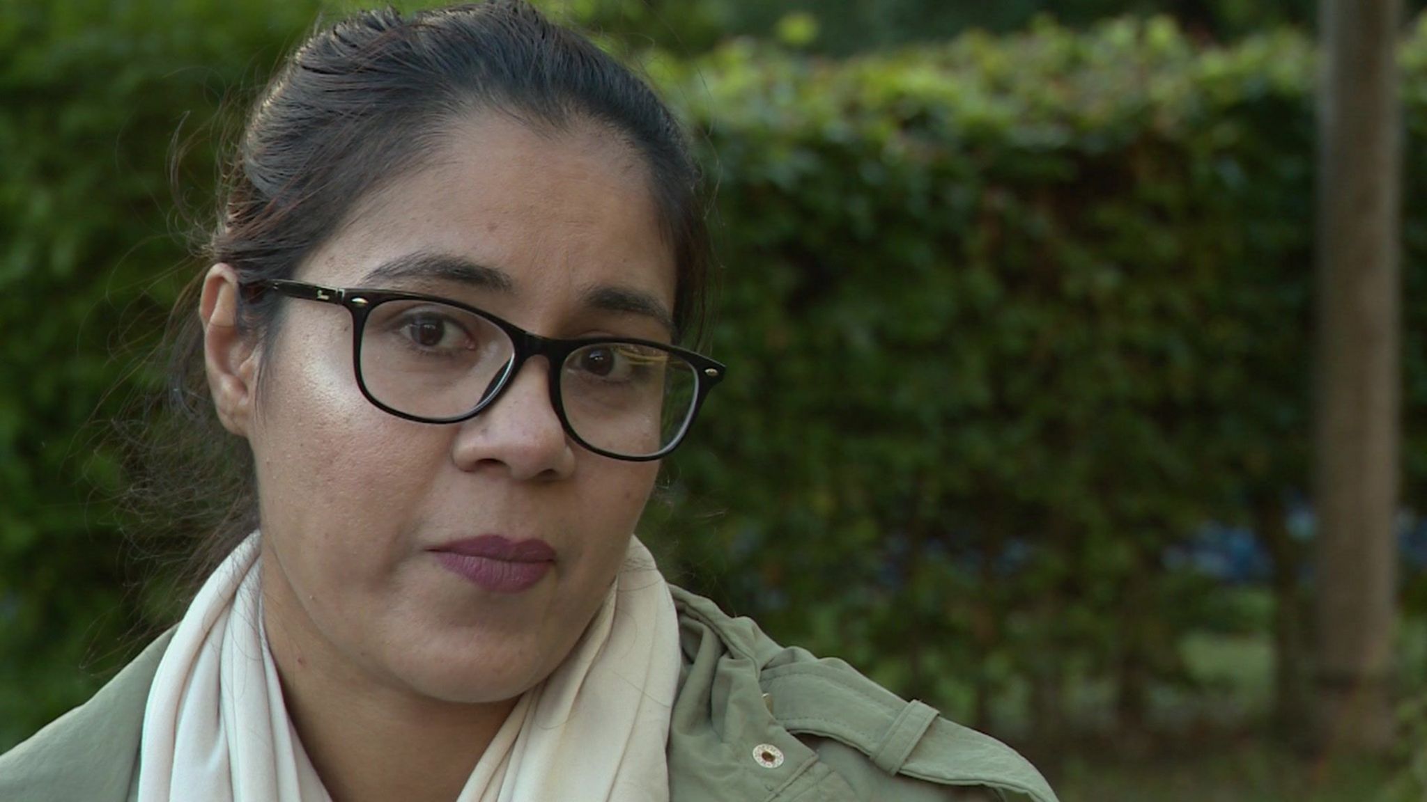 Shahima Kazi, a woman with brown hair and glasses wearing a khaki jacket and cream scarf, sitting in front of a green hedge