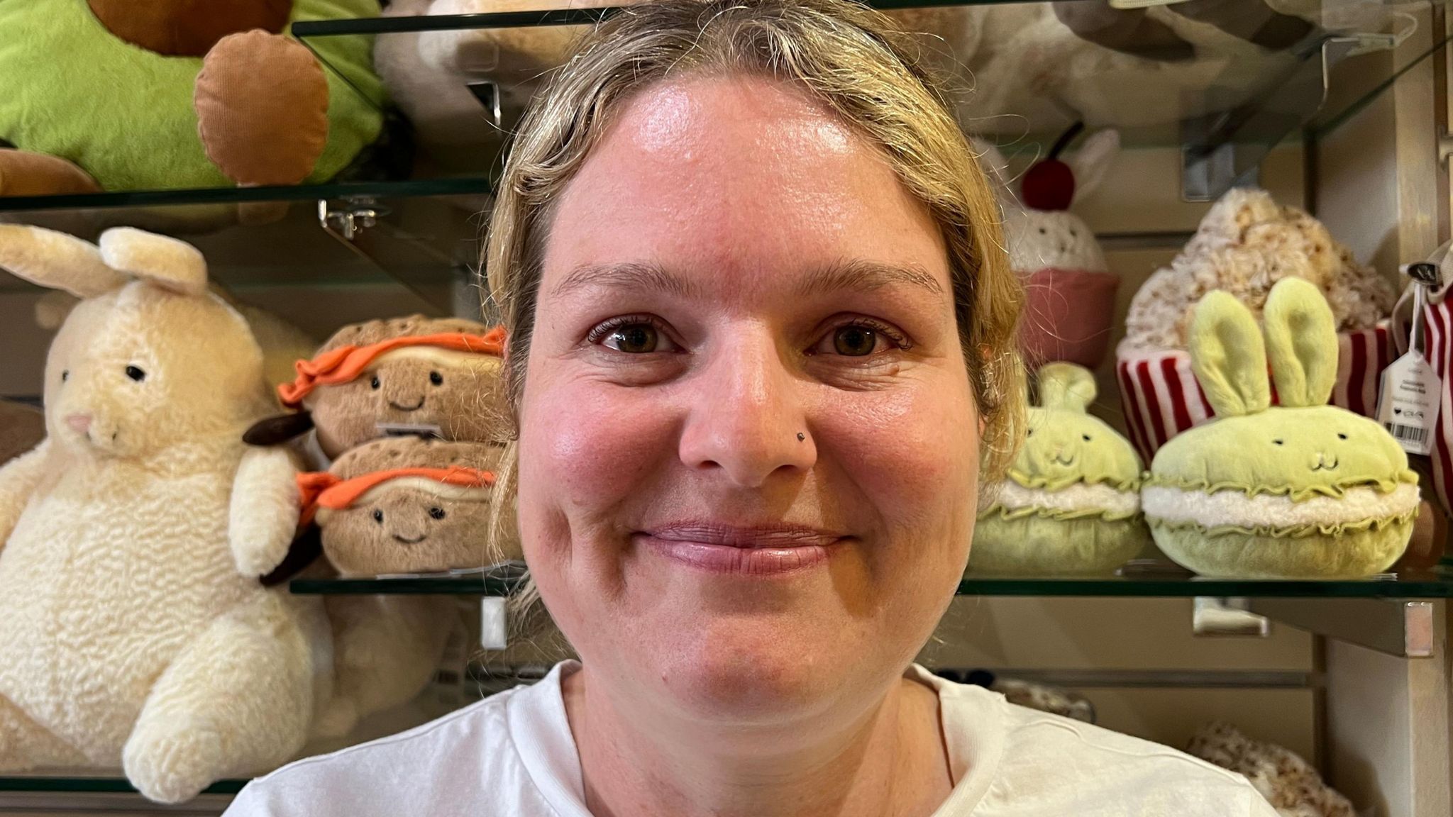 Emma Steele is smiling, she has brown eyes, pink lips and rosey cheeks. She's standing in front of a display of jellycat plush toys. To her right are two green bunnies, to her left is a plush bagel sandwich and a white bunny.