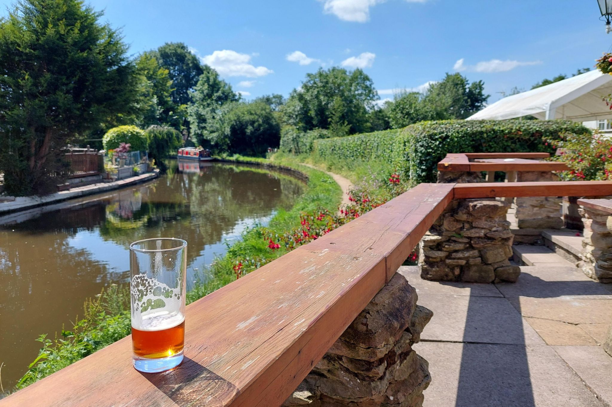 A canalside scene, with a glass of beer sitting on a wooden bench surrounding a patio