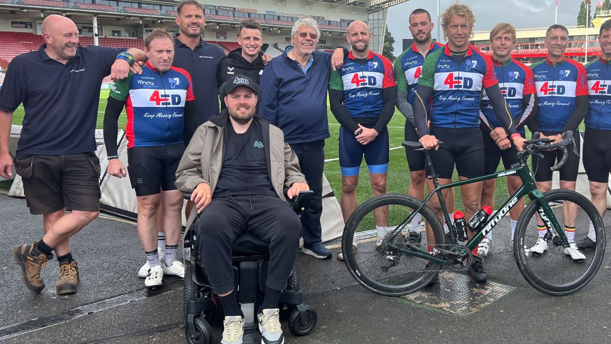 Rugby players who took part in the 24 hour cycle ride line up for a group photo in a stadium, with Ed Slater at the front in a wheelchair.