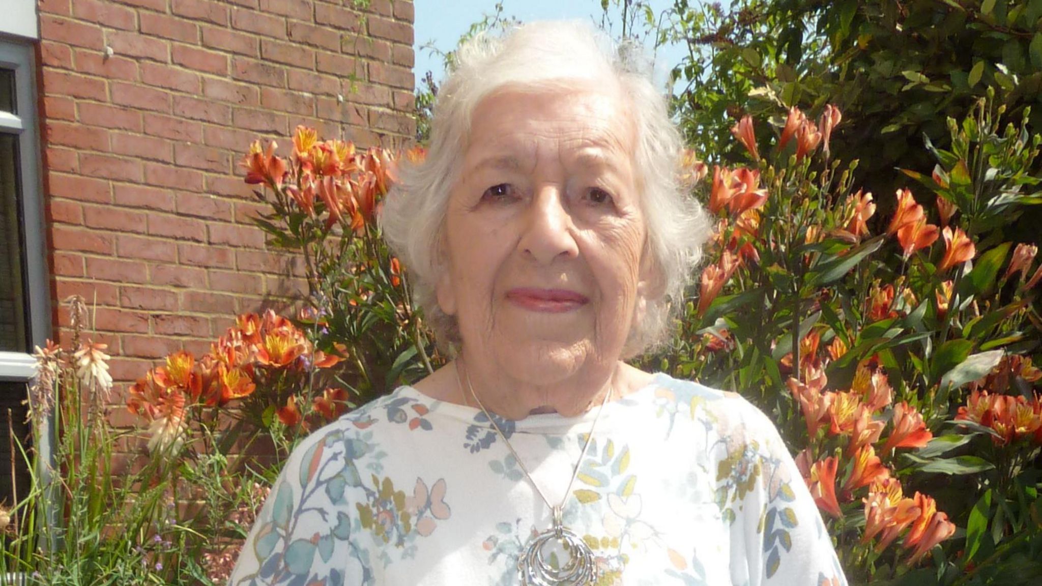 Head and shoulders image of Phyll Babb, wearing a white top and standing in front of some orange flowers