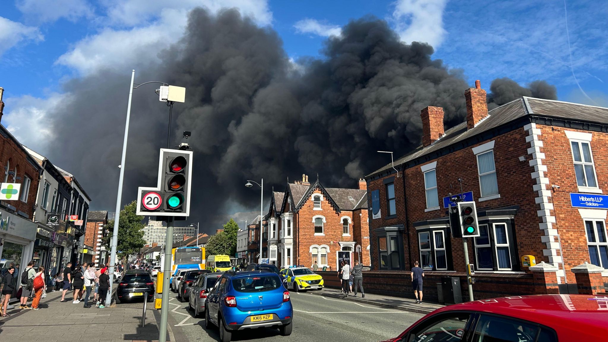 Black smoke billows from behind buildings as traffic builds up on a street with people on the pavement watching on.