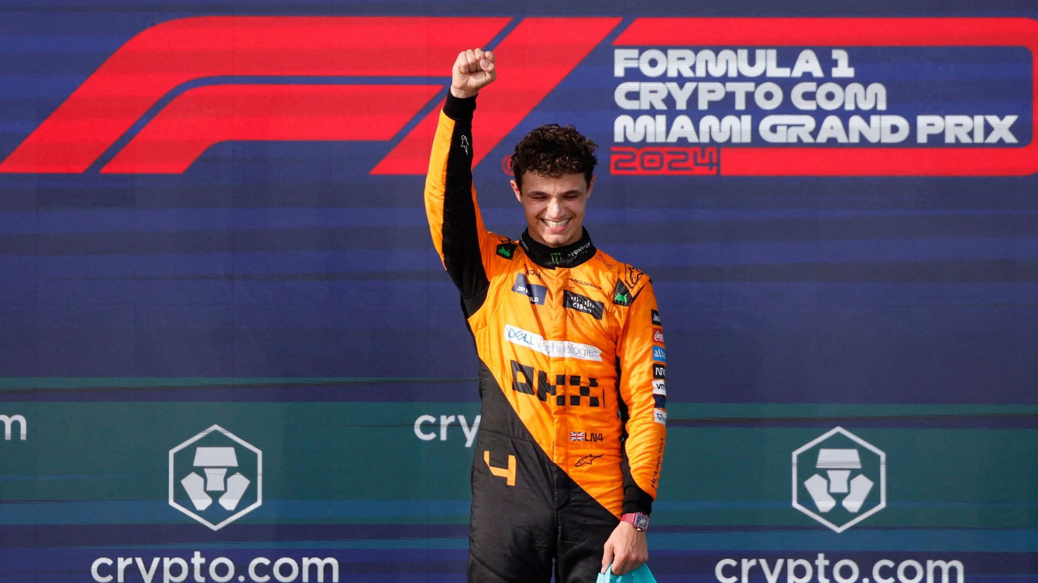 Image of Lando Norris after the Miami Grand Prix. He is wearing an orange and black racing suit, with his fist in the air. His eyes are closed and he is smiling. 