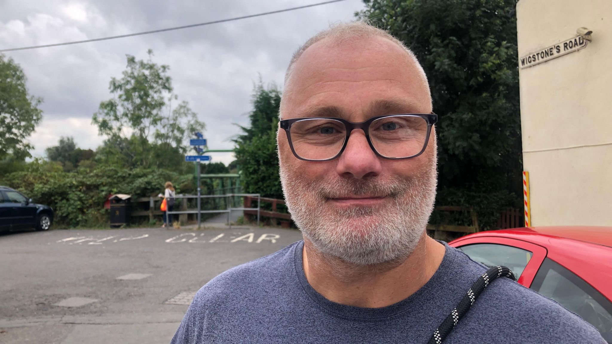 Lee Lambert smiles and looks directly at the camera, he is photographed in a car park and wearing a blue top and black glasses. 