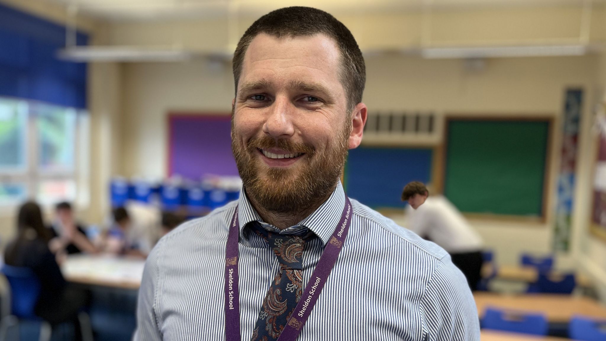Teacher, Richard Page, with short brown hair and bears, a blue striped shirt and multi-coloured tie - smiling