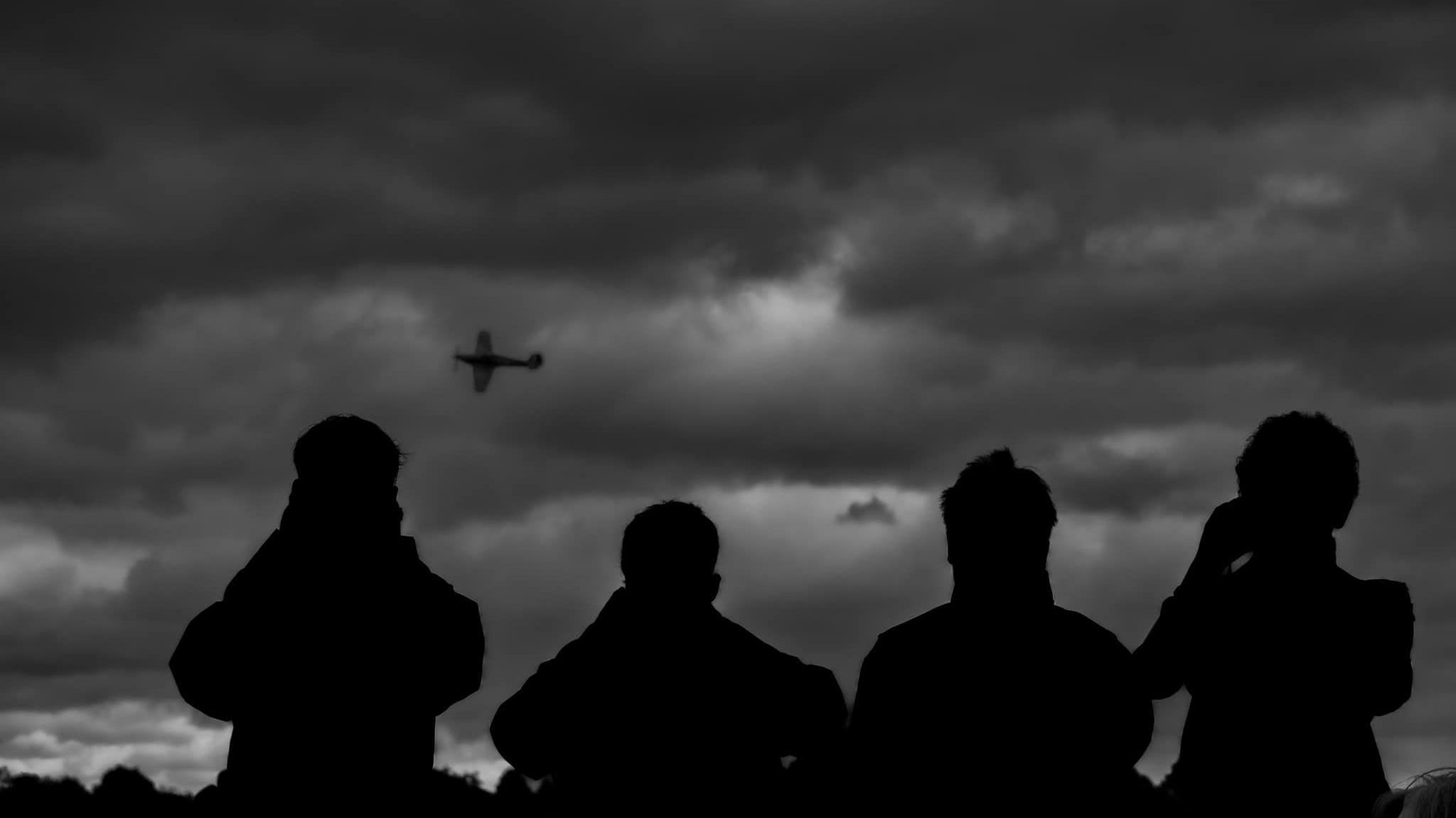 Silhouettes of four people watch the sky, where there is a plane flying in front of them
