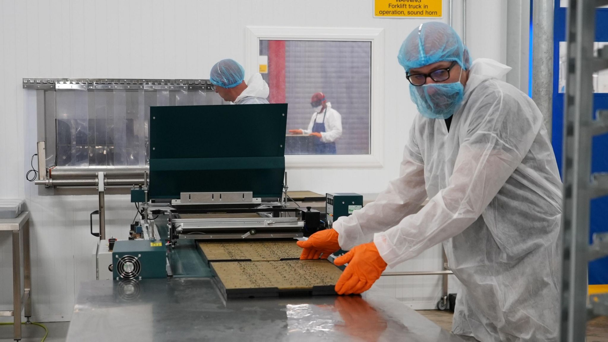 A man in a white plastic suit with a blue mask and hair net and orange gloves moving a tray of seeds with two other people in the background