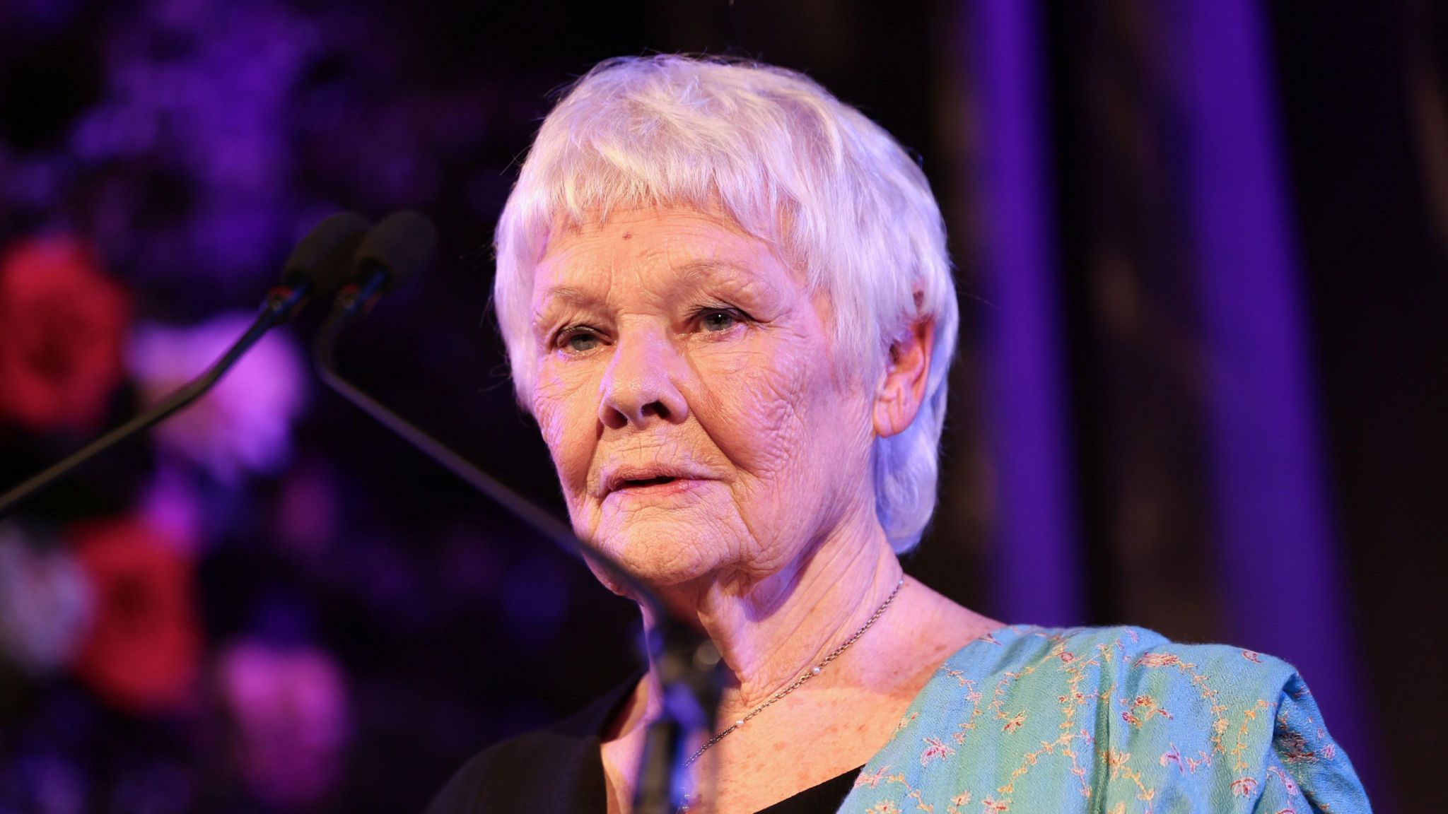 Dame Judi Dench speaking at an event. She is standing at a lectern with microphones and is wearing a green dress.