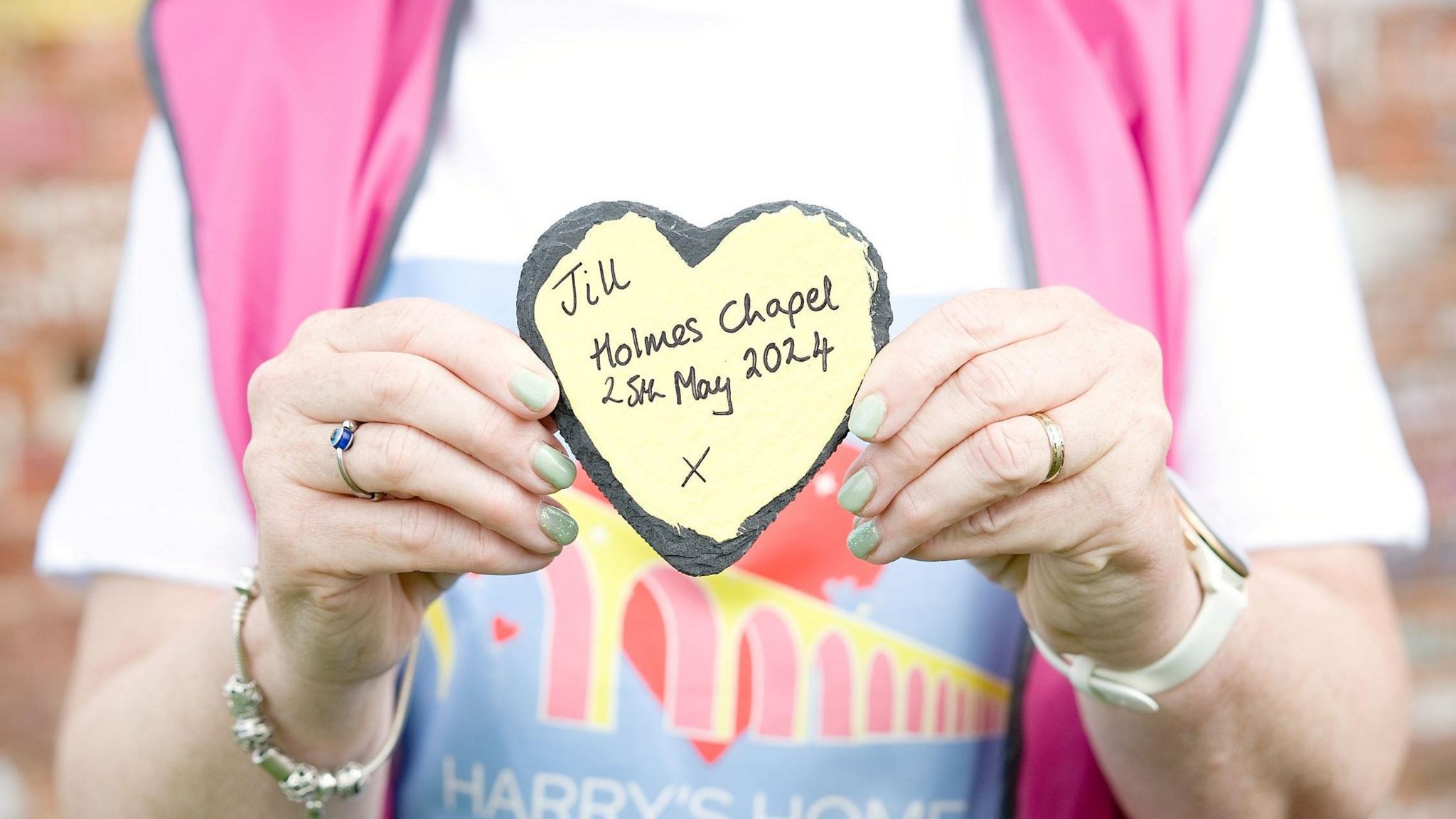 Woman holds stone heart up with the words 'Jill, Holmes Chapel, 25 May' written on it.