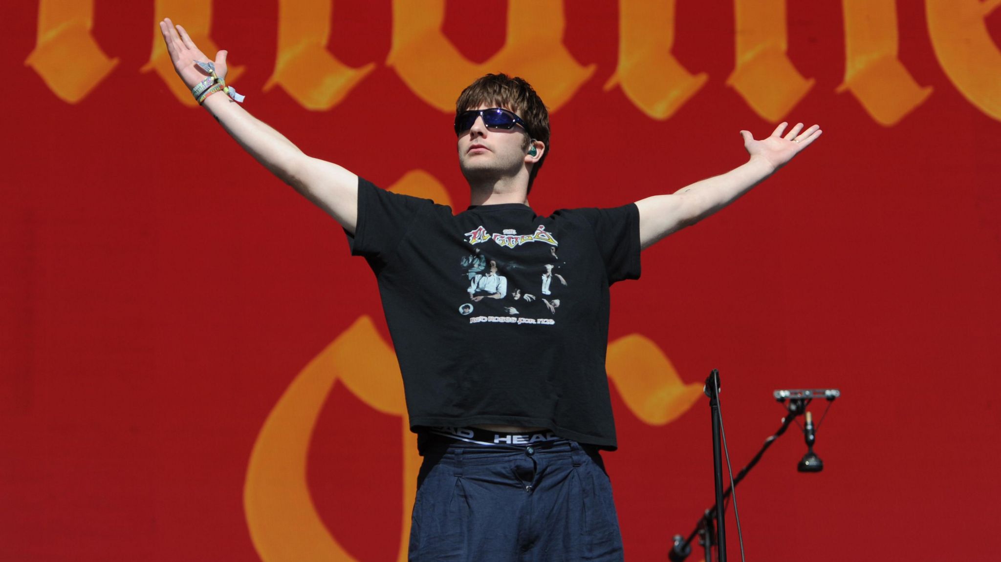 The lead singer of Fontaines D.C., Grian Chatten, pictured performing standing with his arms in the air and sunglasses on.