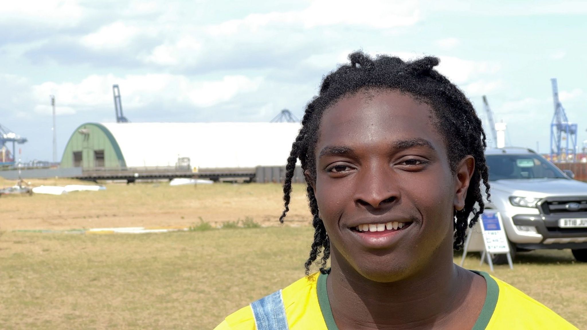 A young man stands in a grassy area. He is wearing a yellow top and is smiling at the camera