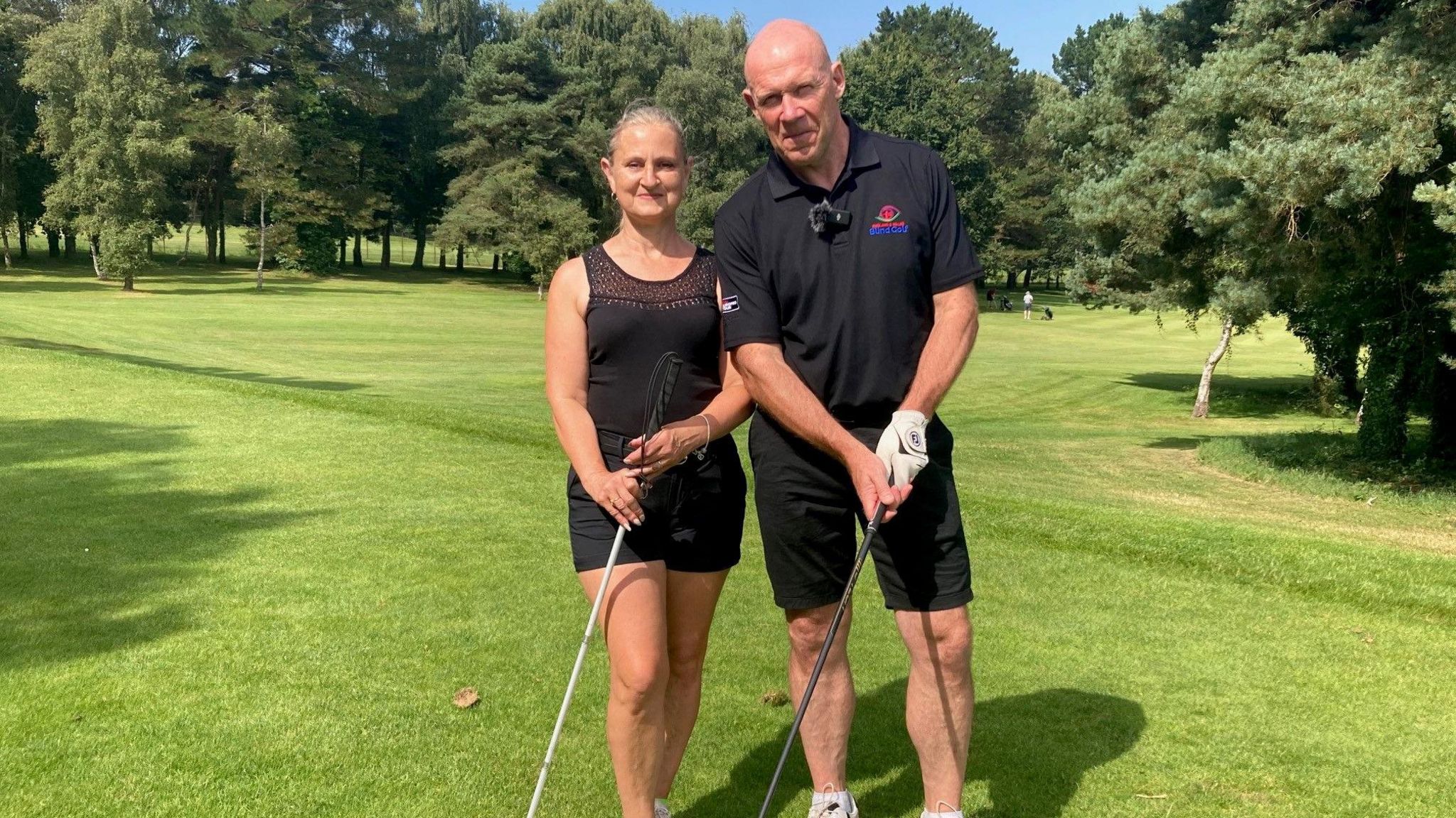 Jane stood next to Simon on a golf course. She is holding a white stick used for lining the golfer up and he is stood with a golf club.