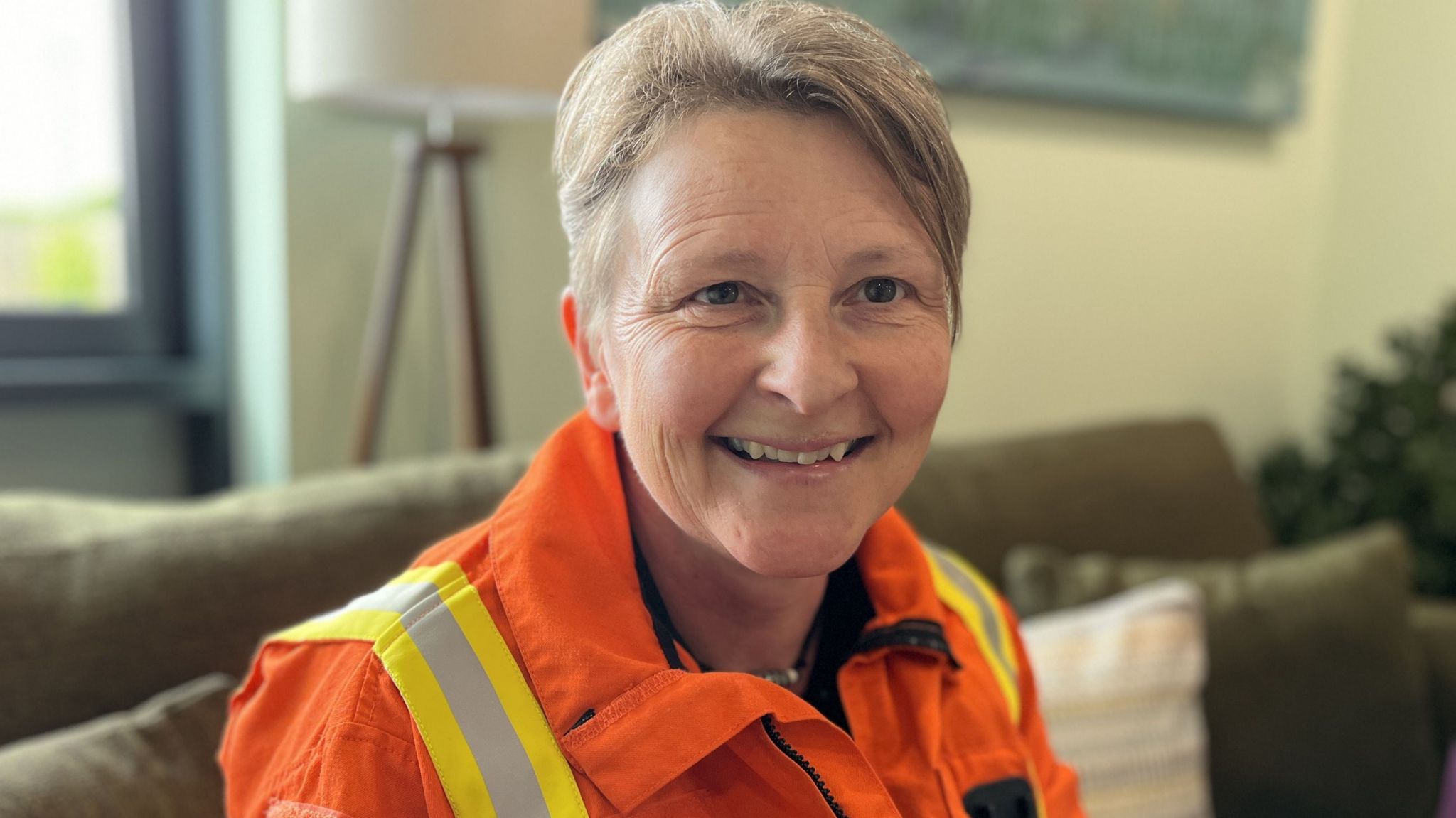 Louise Cox with short mousy brown hair smiling and wearing her orange high visibility uniform sat on the sofa in the charity's family room