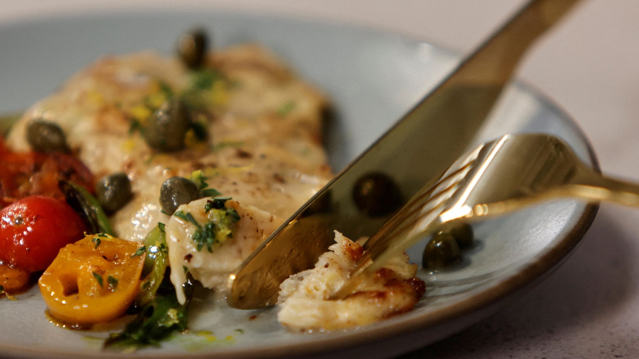 A cooked piece of cultivated chicken breast on a plate alongside some vegetables being cut with a knife and fork.