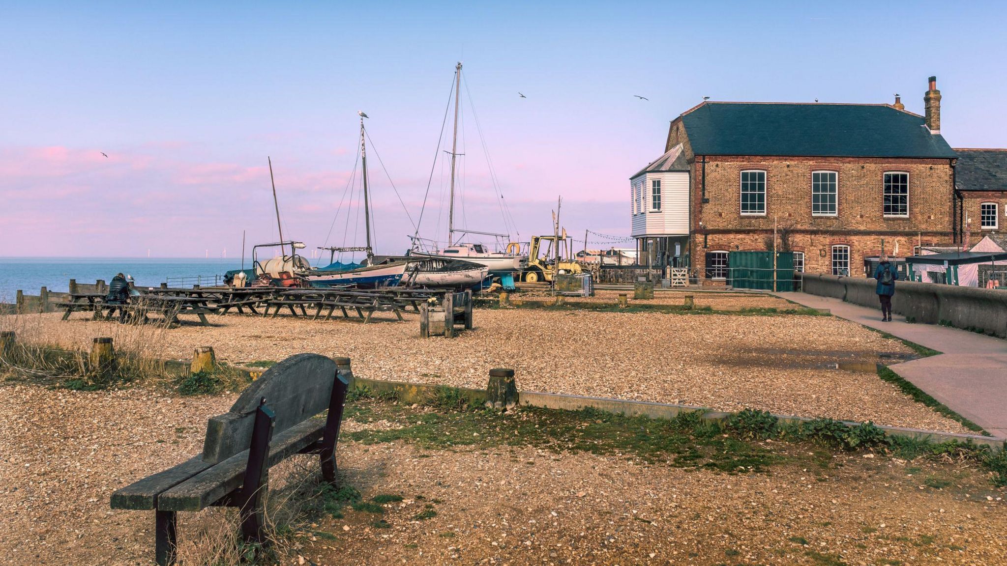Boats pulled upo on a pebble beach behind some benches and tables on Whistable seafront
