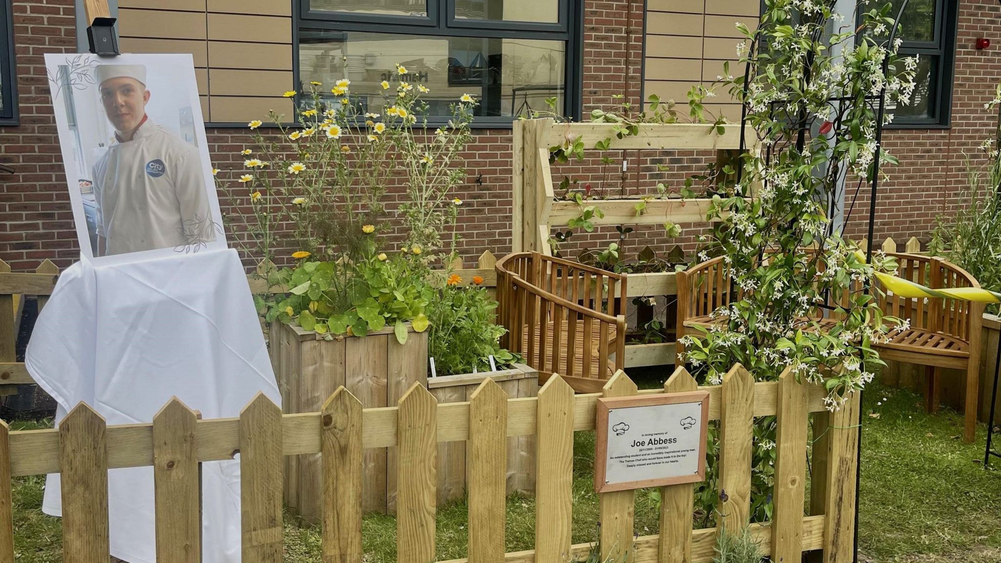 A garden with wooden fencing around and a plaque on the fence which says Joe Abbess on it and there is also an image of Joe printed on card and resting in the garden