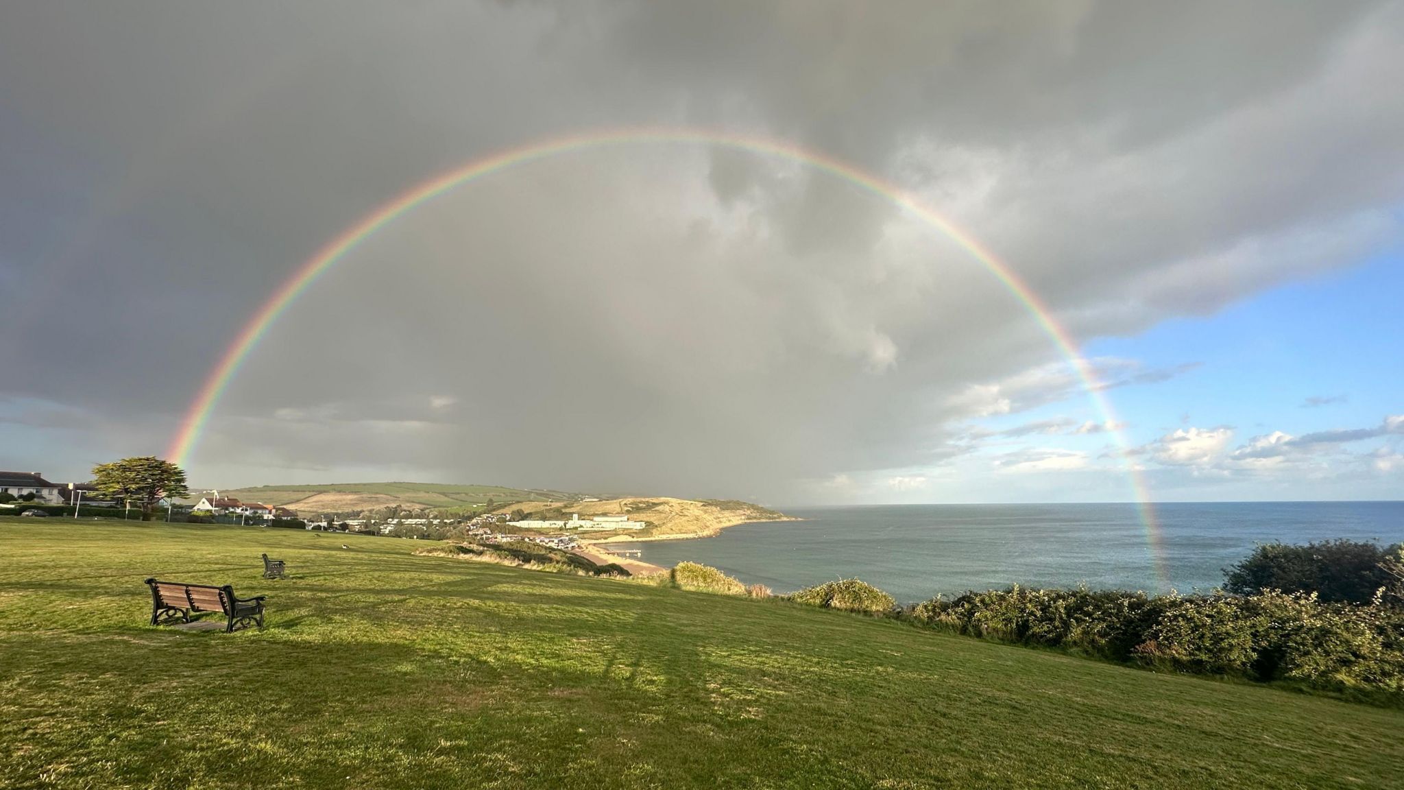 A full rainbow stretches from a behind a tree on a field and over a bay and into the sea under a mixture of dark cloud and bright blue sky in Weymouth