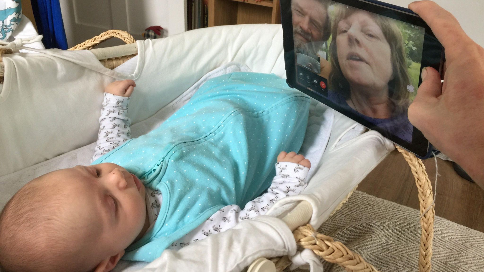 A baby in a blue sleepsuit is lying in a moses basket. Above the basket his father's hand is holding an ipad and on the screen there is a couple in a garden waving hello