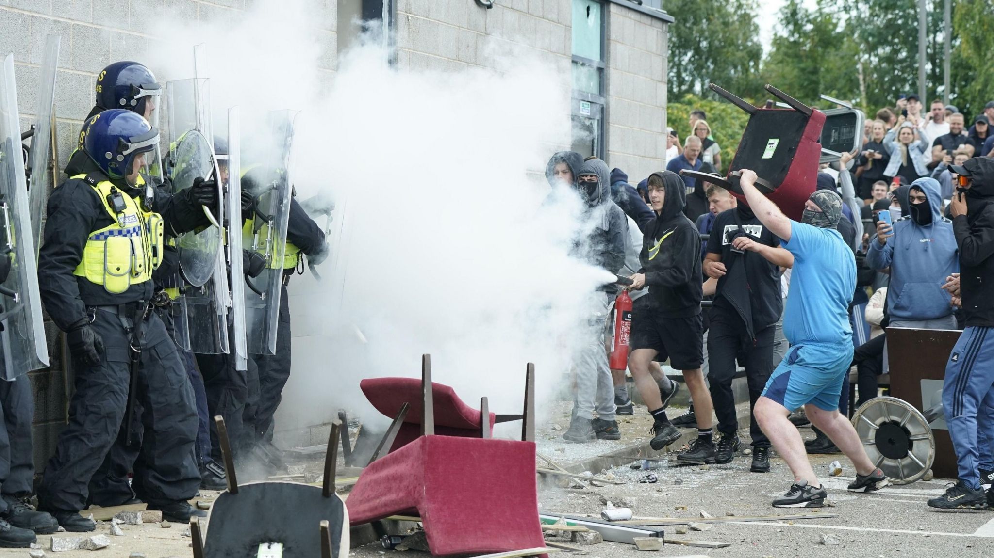 Masked protester holding chair as police officers stand in line next to plume of white smoke