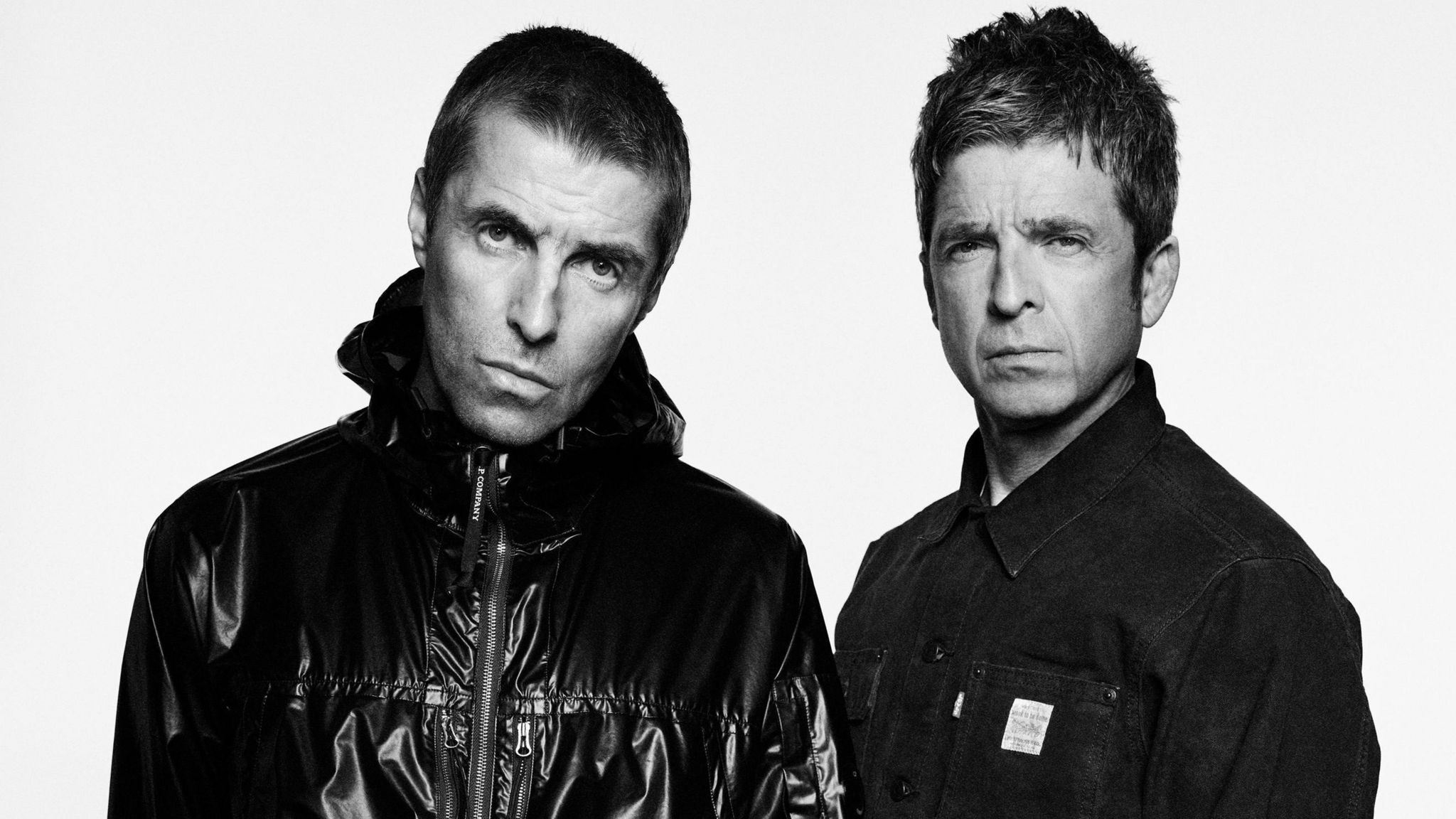 A black and white studio shot of Liam and Noel Gallagher. They are looking moody and staring at the camera