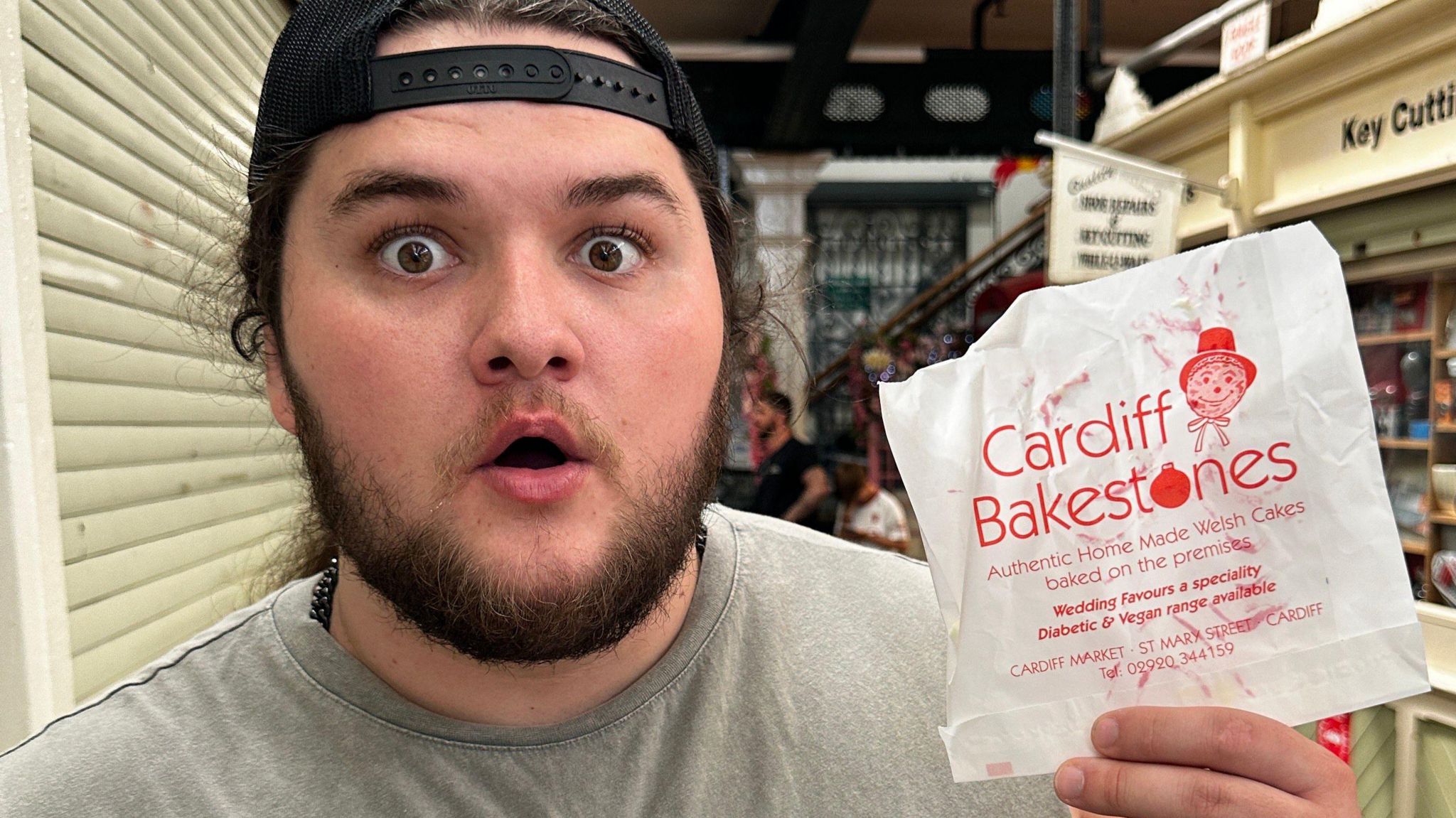 Kalani Smith is a young man with long hair and a beard. He also has brown eyes and is wearing a black mesh baseball cap turned backwards. He is holding up a bag which reads "Cardiff bakestones"