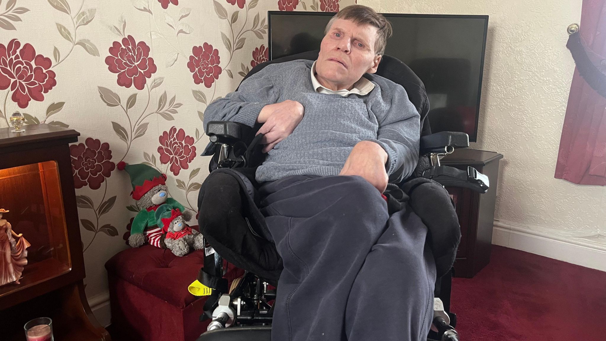 Robert Mason in his wheelchair in his home, with floral wallpaper and a tv in the background