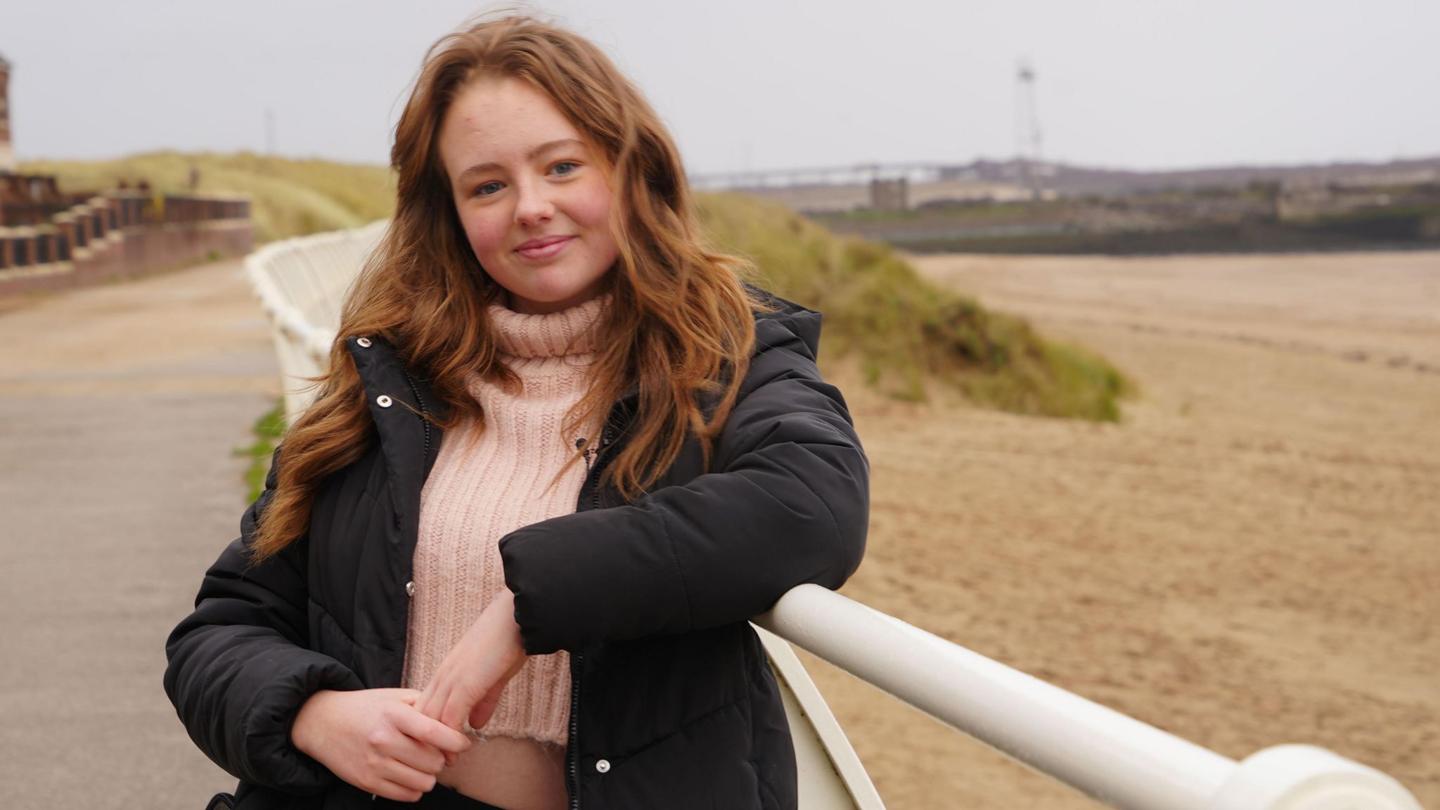 15 year old Lauren stands by Aberavon beach, leaning on a fence, smiling into camera.