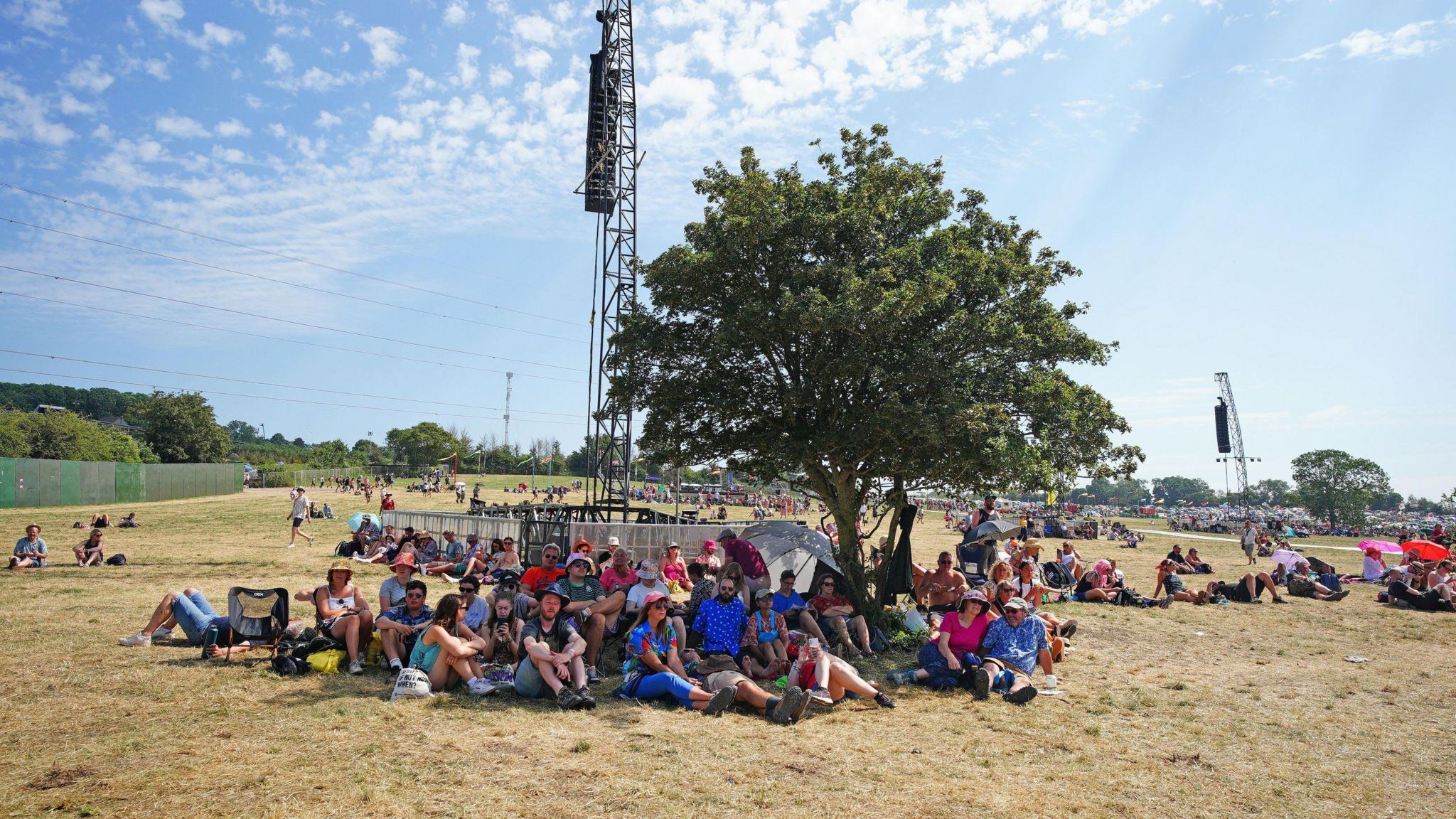 Glastonbury Festival attendees sitting under a tree in the shade on a sunny day