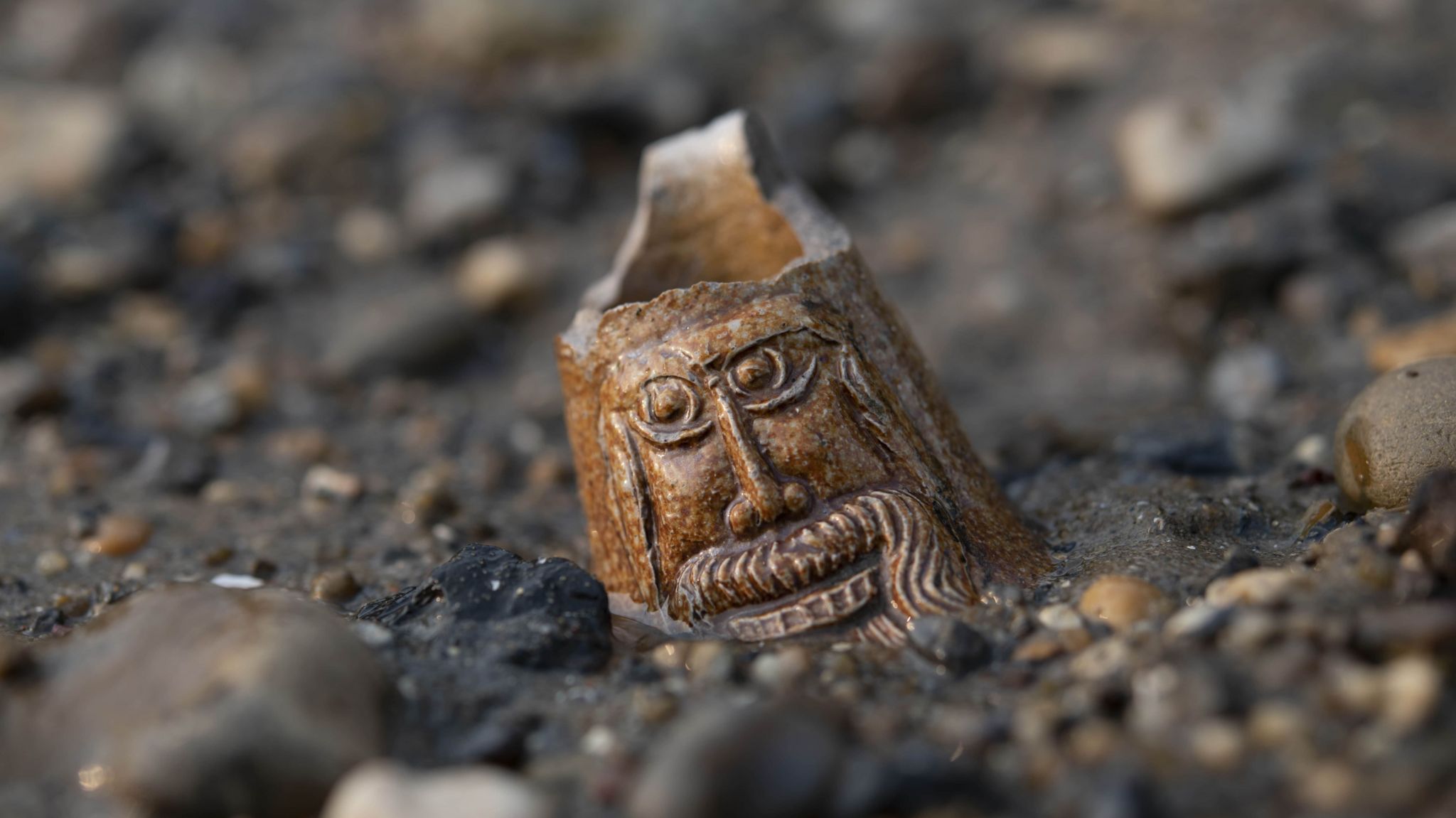 The neck from a stoneware bottle with a bearded face known as a Bartmann bottle from the 1500s to 1600s. The bearded face decorating the neck lies half-buried on the foreshore.