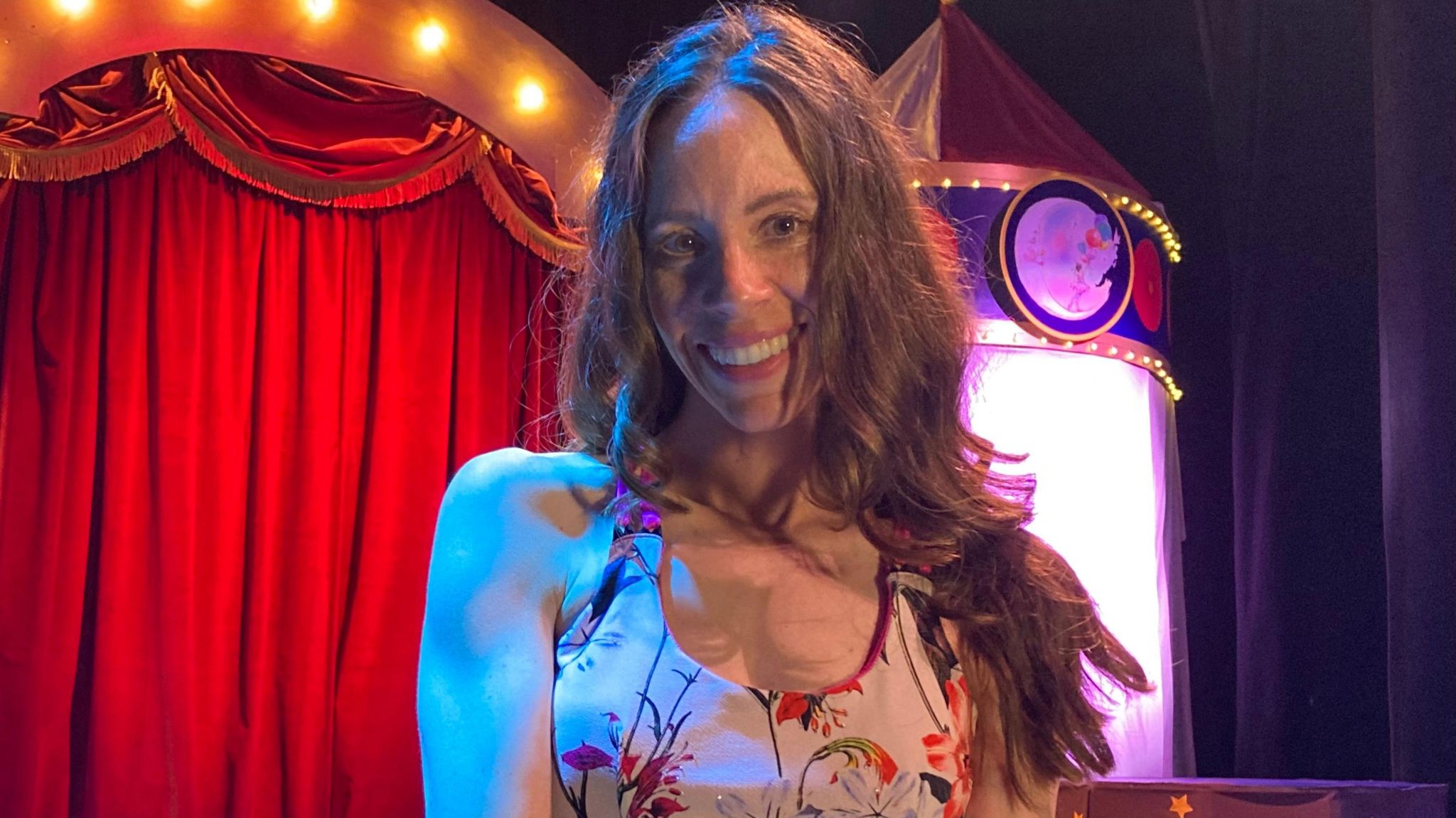 A head and shoulders shot of a woman with long, coloured hair smiling at the camera, in front of a theatre curtain