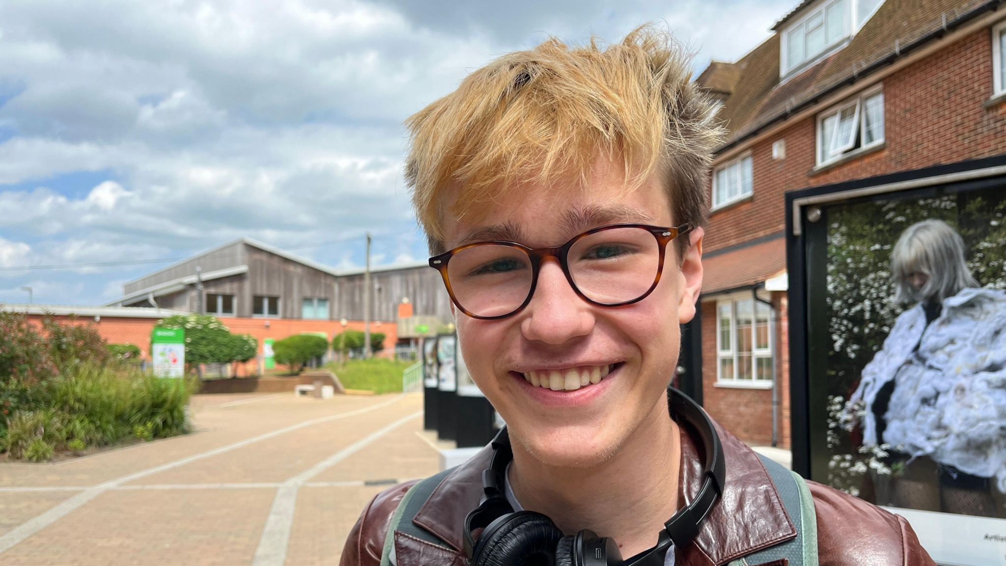 Oliver is 18. He has glasses and bleached blond hair. He's smiling wearing a t-shirt, leather jacket and headphones around this neck.