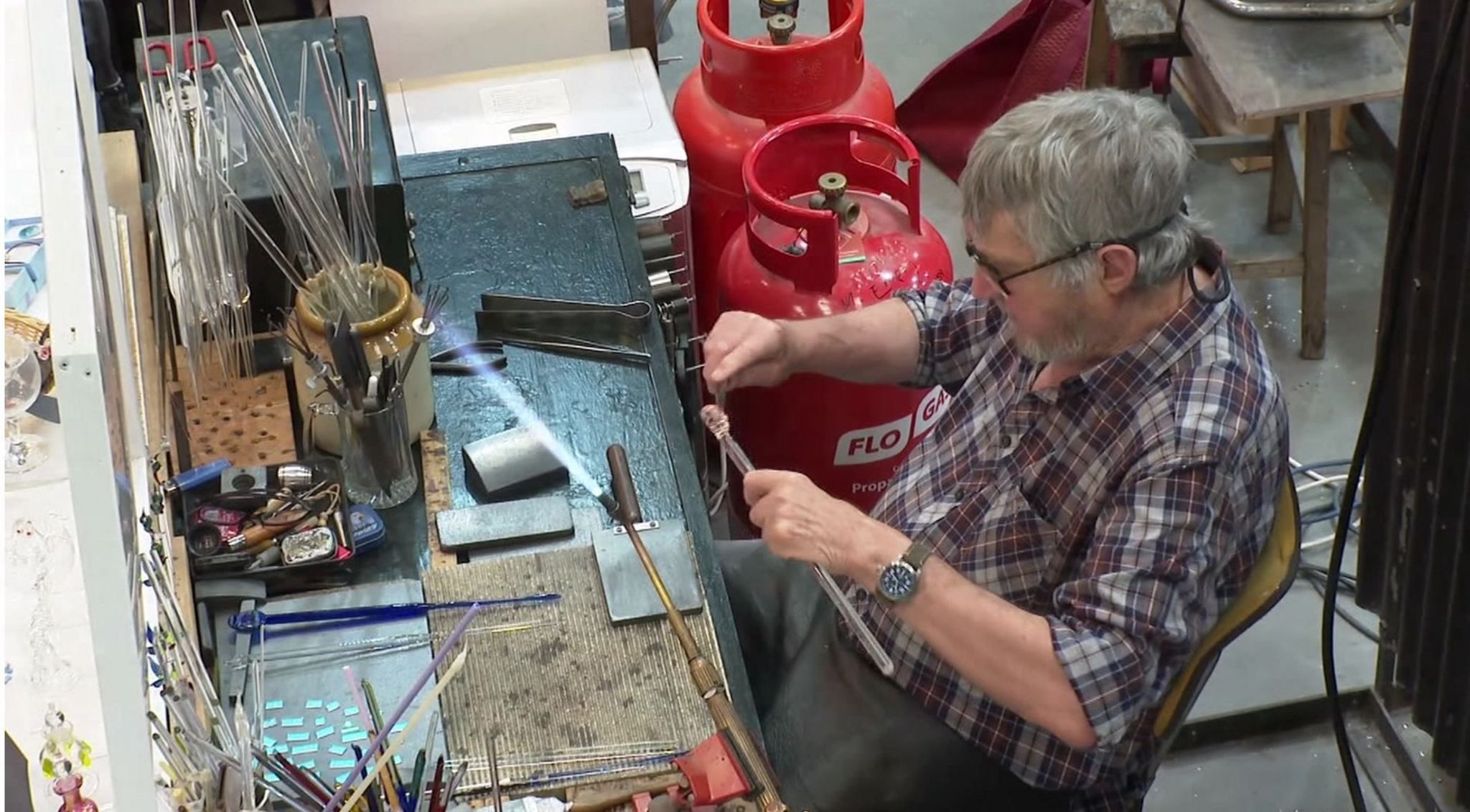 A glass sculptor sitting at a work bench with jars of glass rods, a blowtorch and crafting a glass rod into a sphere. 