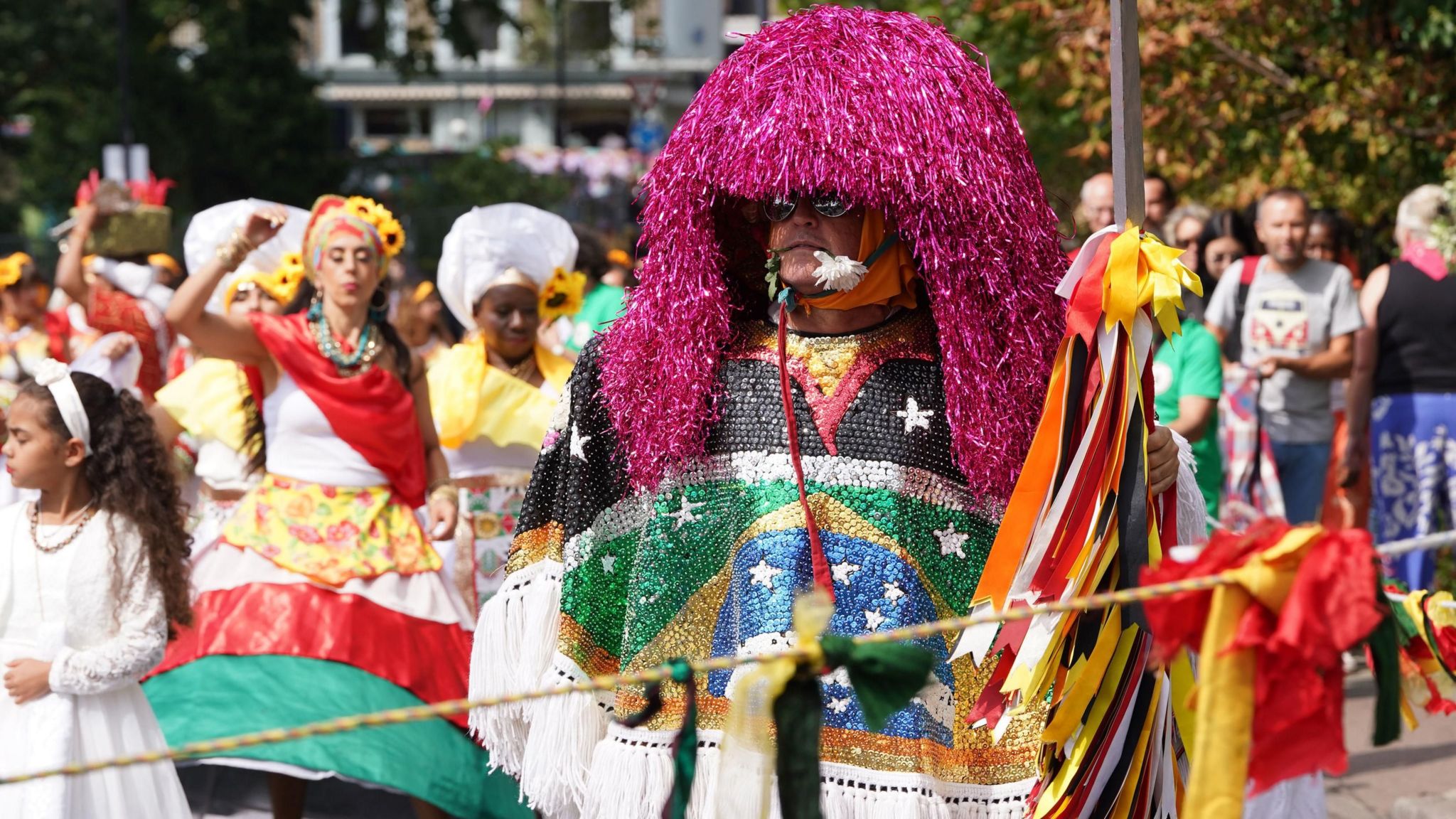 A man dresses up with colourful sequins which make up a Brazilian flag. He's also wearing a purple-streamed headdress and has a white flower in his mouth.