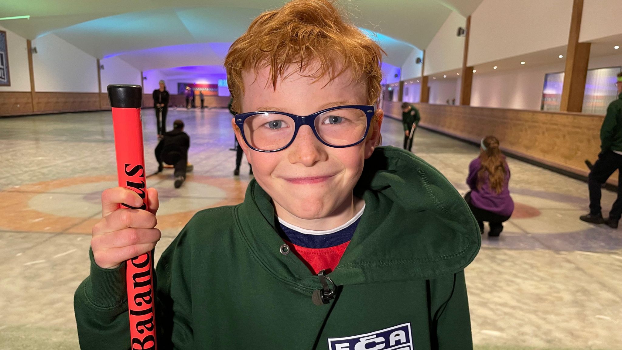 Henry smiles at the camera, wearing dark framed glasses and a green England Curling Association hoodie, with the ECA logo featuring the English flag, plus a top with a red, blue and white collar. He is holding a red curling brush. There are several people curling on the ice rink behind him.