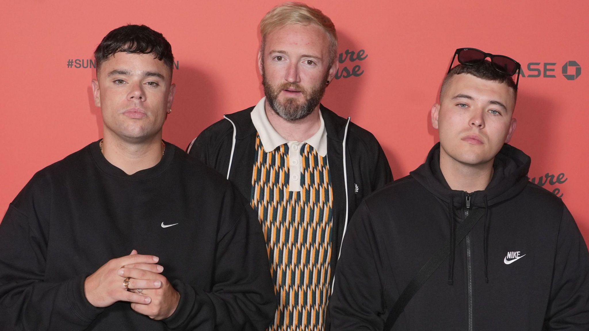 The three band members looking at the camera. One has sunglasses on his head with a dark hoodie. One is wearing a ring and a dark jumper. The other member stnds behind the first two and is wearing a colourful patterned top