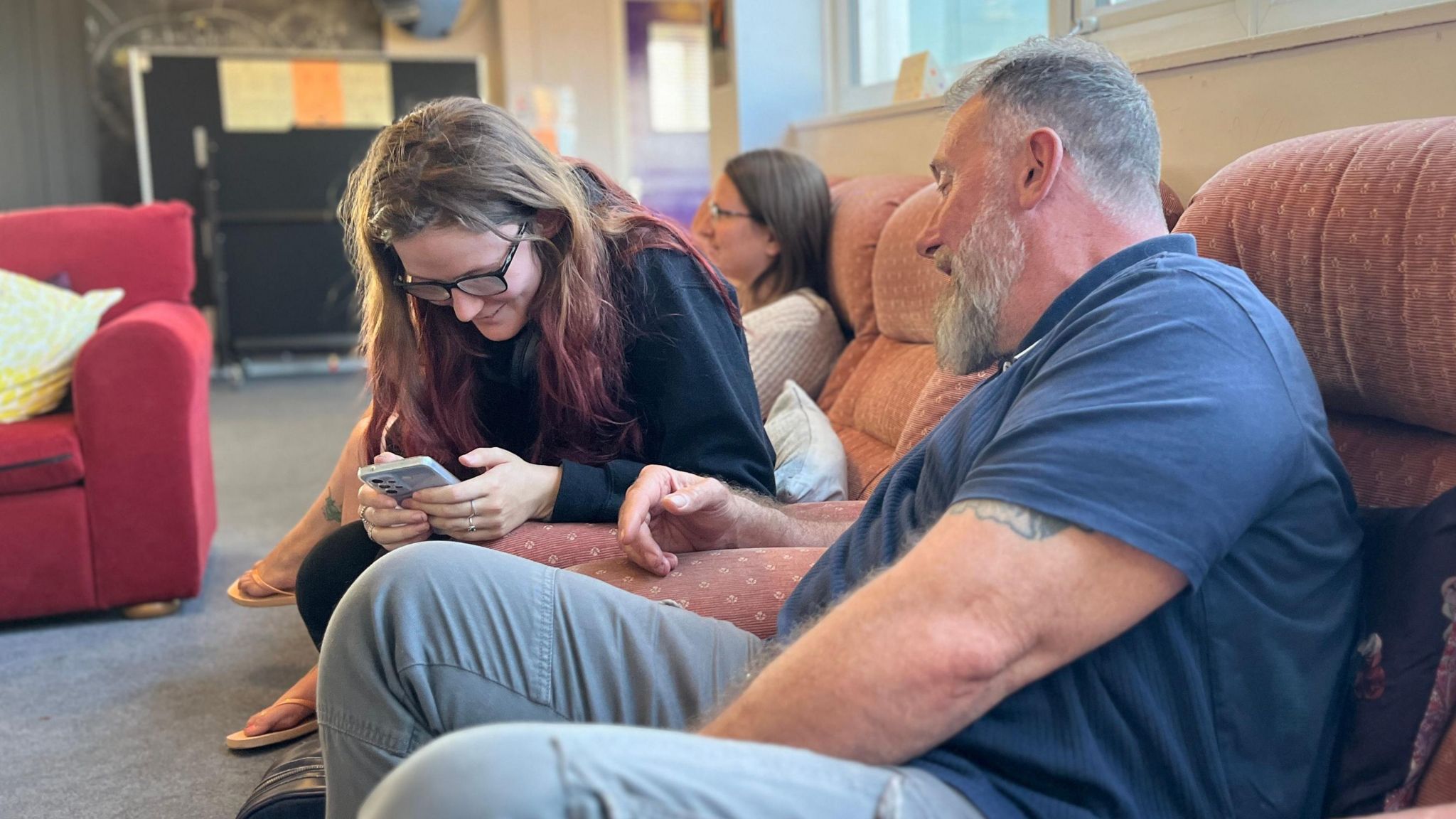 A young women looks at a phone, with an older man sat alongside her on a sofa