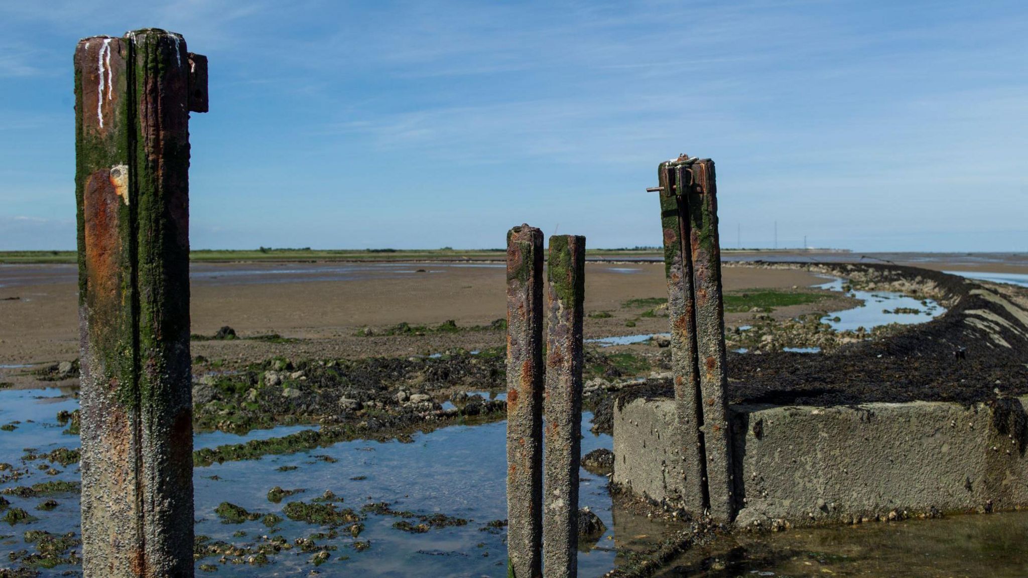 Foulness Island. In the foreground there are rusted pillars, which are the remains of an old oil tank, and sea water. In the background there is shingle and clusters of seaweed and rocks