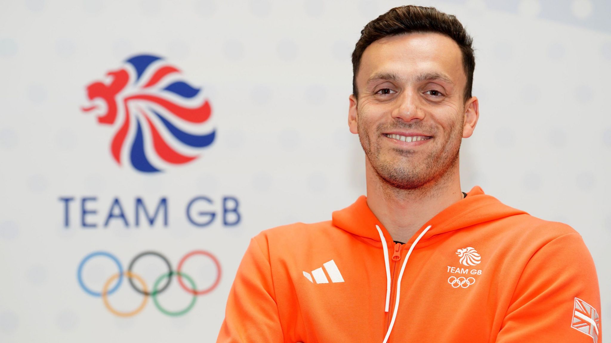 Image of James Guy MBE. He is pictured in front of an Olympic ring sign, with Team GB printed above it. He is wearing an orange hoodie with Team GB logos.